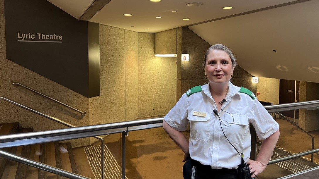 On the left, a sign reads "Lyric Theatre" above a descending staircase with metal handrails, bordered by beige walls. On the right, a woman in a white shirt, dark trousers, and a green vest stands, centered, with a name tag. The floor is beige, and the ceiling, which features recessed lighting, matches the walls in colour and texture.