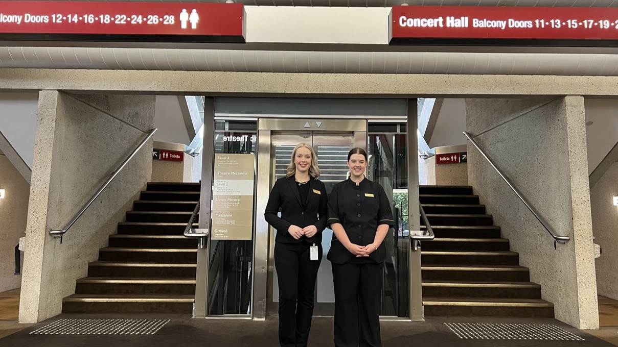 On both sides, wide staircases with handrails and truncated domes rise symmetrically, alternating between dark and light steps. In the middle, a lift is positioned beneath a digital sign displaying numbers for balcony doors. Two women dressed in formal uniforms stand in the foreground. The floor is covered in grey carpet, while the ceiling is obscured, drawing attention to a prominent interior structure leading to the entrances.