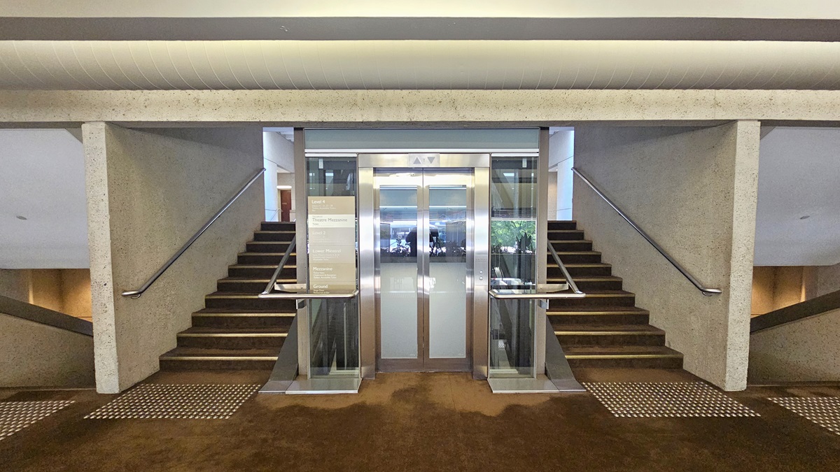 In the middle, there is a lift equipped with a poster. Stairs with handrails and truncated domes are installed on both sides of the lift. Another set of descending stairs with handrails and truncated domes is also installed on both sides of the stairs. The ceiling and wall are made of concrete and have air conditioning vents.