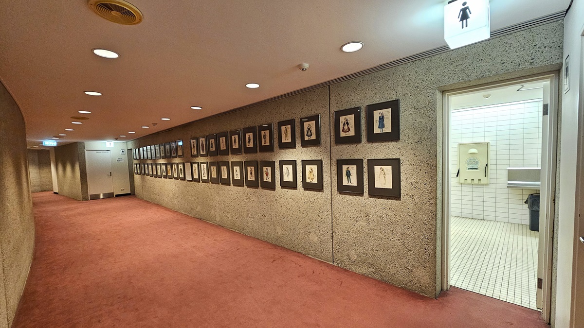 A corridor with pink carpet leads to the regular female toilets. A "female" sign is affixed to the ceiling near the door. Inside the toilet, a foldable change table is mounted to the wall. The wall and floor have white tiles. Outside the toilet, the ceiling is white with LED lights. Framed posters line the concrete wall on the right.