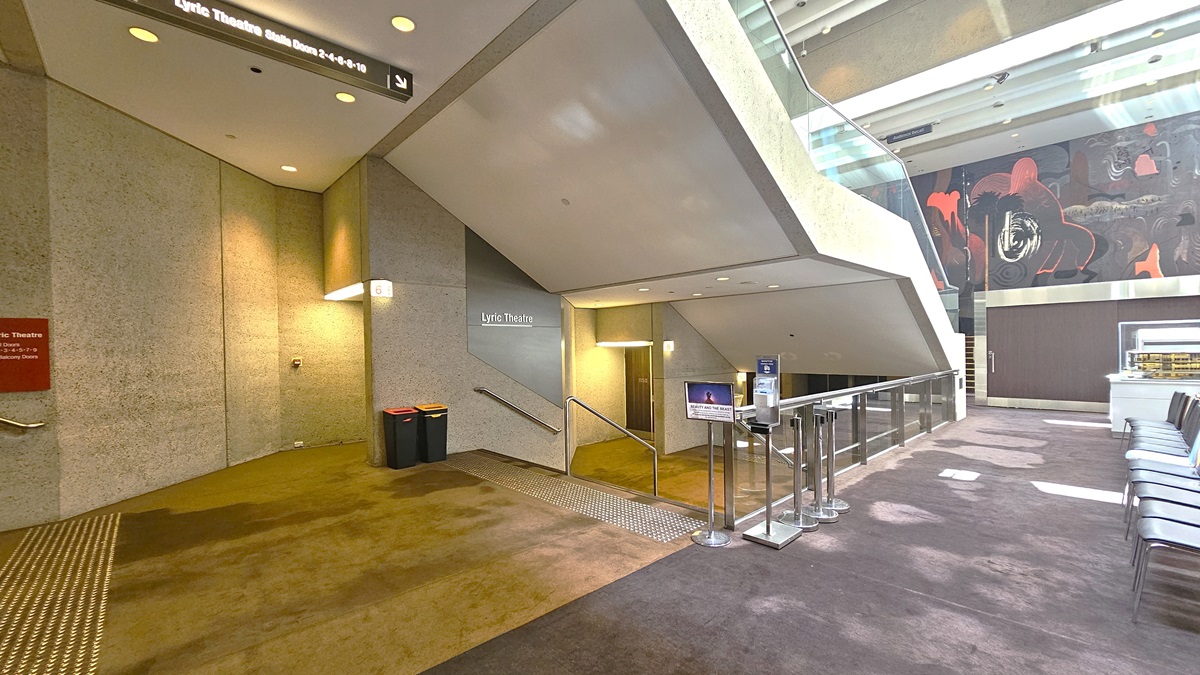 A spacious lobby with beige walls and symmetrical staircases on either side. A large abstract mural at the far end. In the centre, a low table with brochures, and movable chairs arranged side by side, flanked by rope bollards. Bright overhead lighting illuminates. Stairs with handrails and truncated domes are installed on both sides, along with signages. The floor is carpeted.