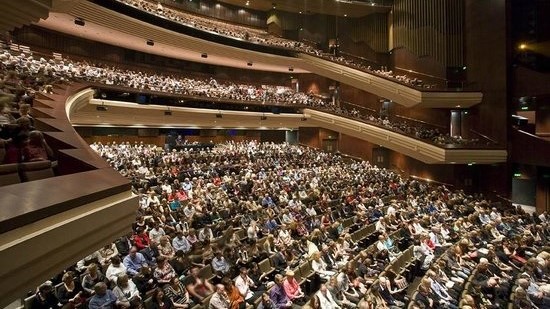 A panoramic view of the fully occupied concert hall with seating arranged on the first, second, and third levels. The wall features a wooden structure.