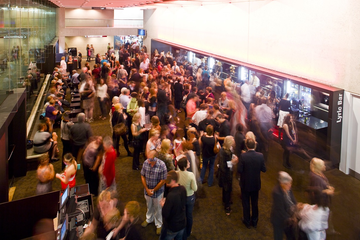 An overhead view of a lobby furnished with wooden tables and bench chairs for seating. On the right, the Lyric Bar is visible, with a lengthy bar counter and refrigerators stocked with cold beverages. A reception desk with computers is situated on the left. The floor is covered in brown carpeting, while the ceiling is pink and adorned with light fixtures. The area is currently crowded with guests.