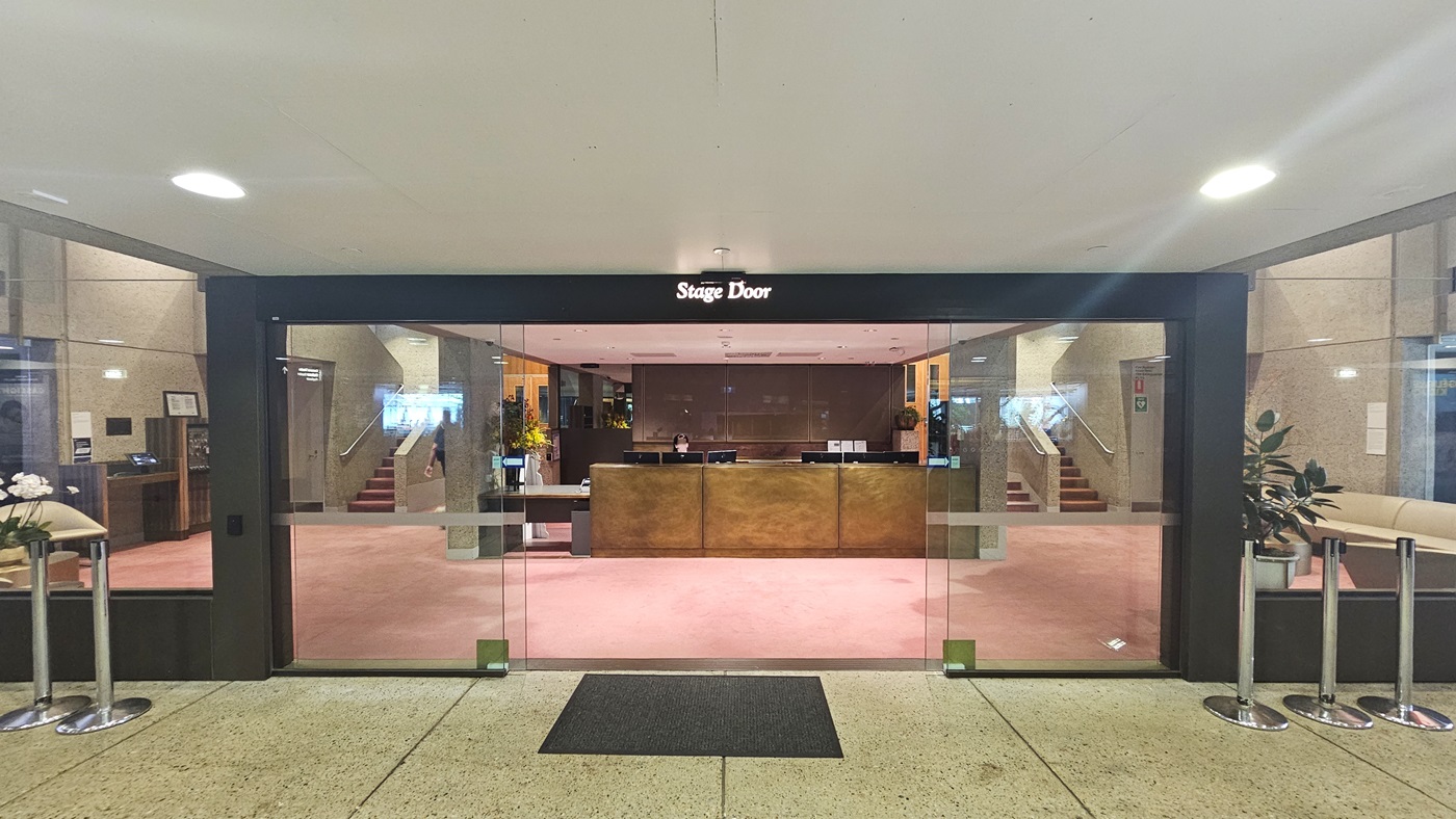 An automatic glass sliding door equipped with a non-slippery mat welcomes visitors. Ribbon bollards are lined on both sides for added safety. A signage reading "Stage Door" is prominently displayed on top of the glass door. Upon entering, the reception desk with a wooden front is located directly ahead. Stairs with handrails on both sides provide access to different levels. The floor features pink tiles, while the ceiling is painted white.