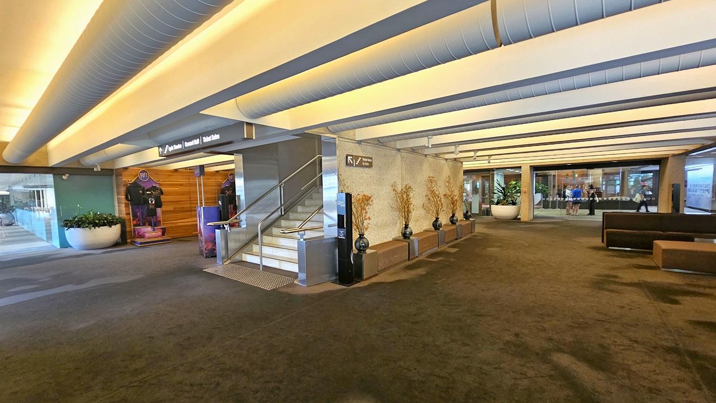 On the left, there's a retail shop displaying T-shirts. Adjacent is a staircase with handrails, equipped with truncated domes. Next to it, there's a line of vase decorations against the middle wall. To the right, chairs are arranged. Further in, more retail shops. The floor has carpet and the white ceiling has large aircon vents. Indoor plants in huge pots are also visible.