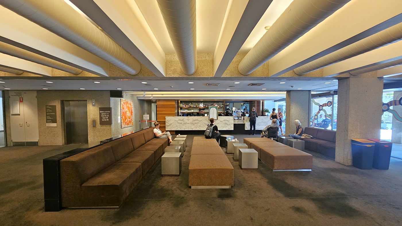 To the right, a sitting area furnished with lounge chairs and coffee tables. Adjacent, a bar counter displays wines, liquors, and drinks, featuring a marble design front. A lift positioned to the left side, equipped with directional signages. A large painting hangs on the middle wall. The floor has dark grey carpet, and the ceiling features white with large aircon vents and LED lights. Guests are visible in the area.