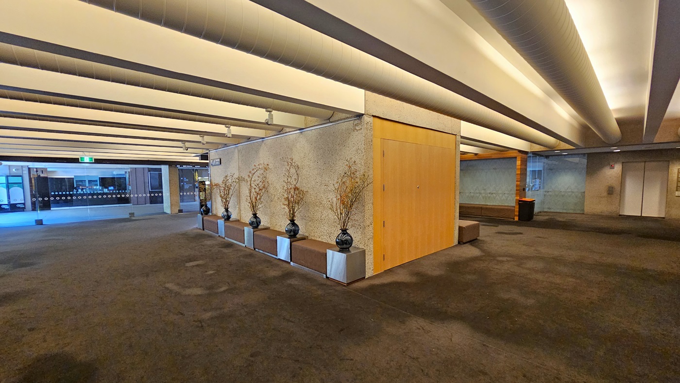 On the left, a glass entrance. In the middle, a line of vase decorations against the wall. To the right, chairs are arranged, with rubbish bins against the wall. The floor is carpeted, and the white ceiling has large aircon vents.