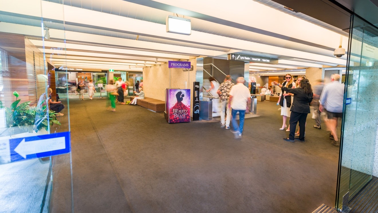 A glass sliding door, marked with blue directional signage, reveals a hallway. People are scattered within, some gathered around a prominent advertisement. To the right, more individuals converse. In the middle, a staircase with handrails stands adjacent to a seating area. Additional seating is positioned on the far right. The floor boasts a brown carpet, while strip lights adorn the ceiling. Indoor plants add a touch of greenery.