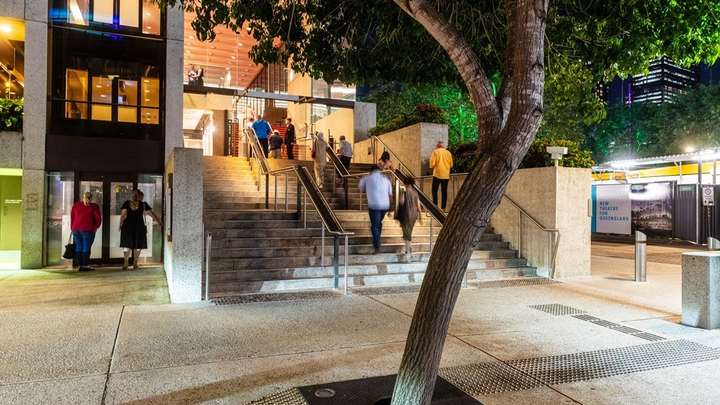 To the left, a group of people stands beneath illuminated building eaves. On the right, a flight of steps with handrails leads to another well-lit area with more individuals. Truncated domes are installed before the stairs. A mature tree with dense foliage sits centrally. The foreground showcases textured paving and truncated domes. A construction barricade is visible on the far right.