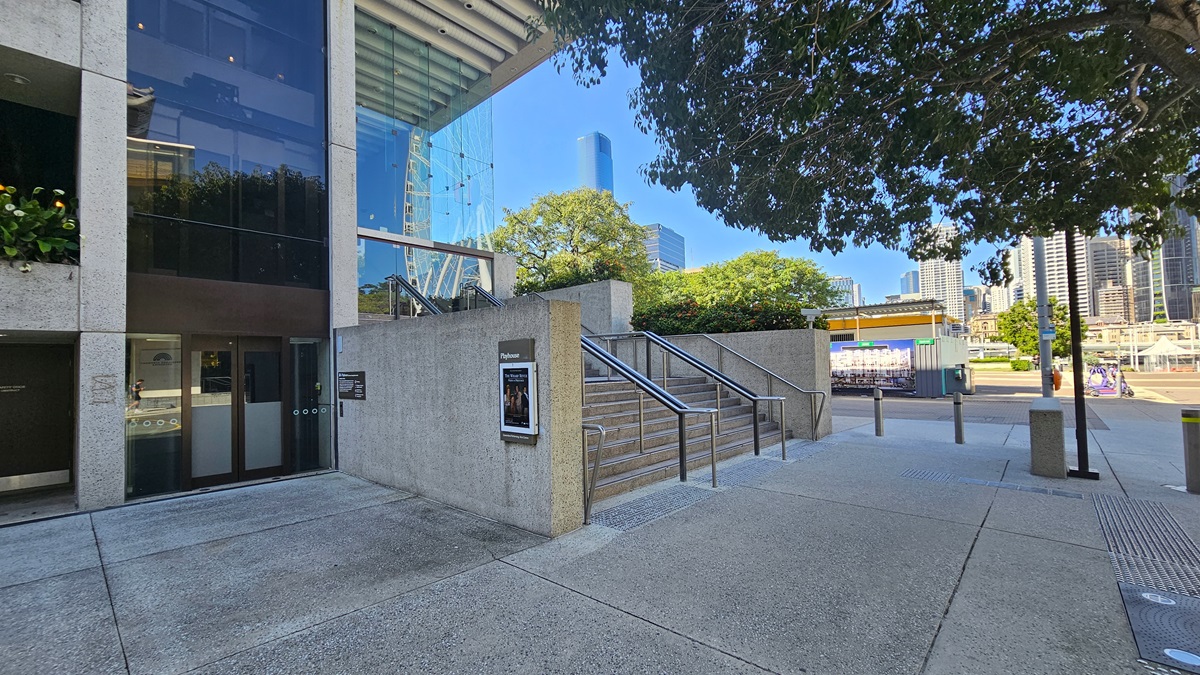 A modern building on the left, featuring a concrete facade and angular design equipped with stairs with handrails. ornamental landscape adorns the front. A metal pole with multiple signs stood beside a solar street light. Rubbish bins are visible. To the right, there is a walkway with trees providing shade and a blue construction barrier on the right.