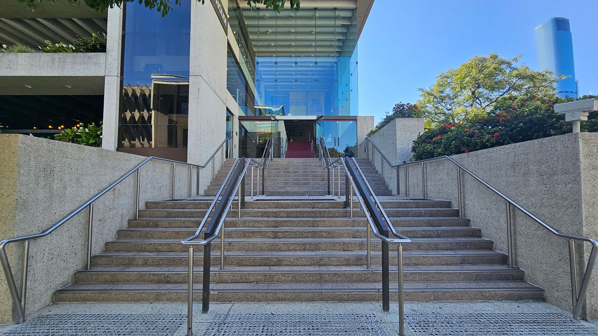 A multi-storey building with a concrete and glass exterior. Stairs with handrails and truncated domes lead to an automatic glass sliding door entrance. On the right, a landscape of plants in concrete planter boxes. Further ahead, tall buildings and trees are visible.