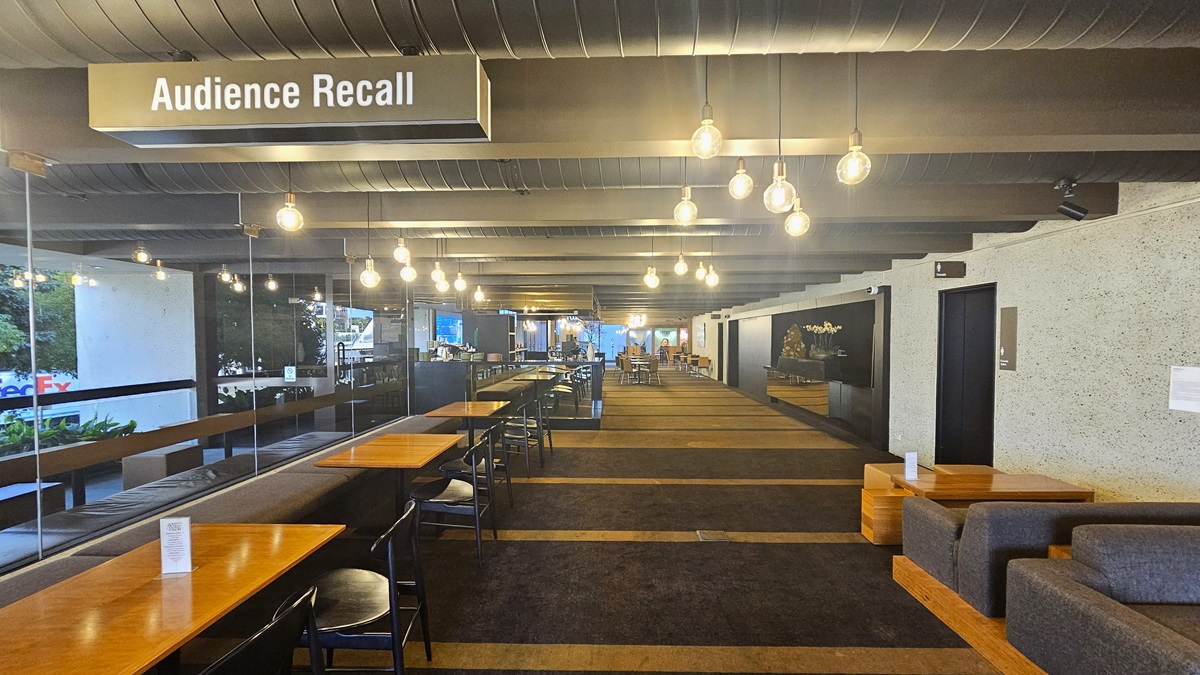 A carpeted area with rows of tables and movable chairs, along with cushioned bench chairs positioned on the left. On the right, couches and coffee tables are arranged against the wall. Further inside, more seating. Brown carpeted floor, with a dotted white wall on the right and a glass wall on the left. The ceiling features large aircon vents and light fixtures. A sign reading "Audience Recall" hangs from the ceiling.