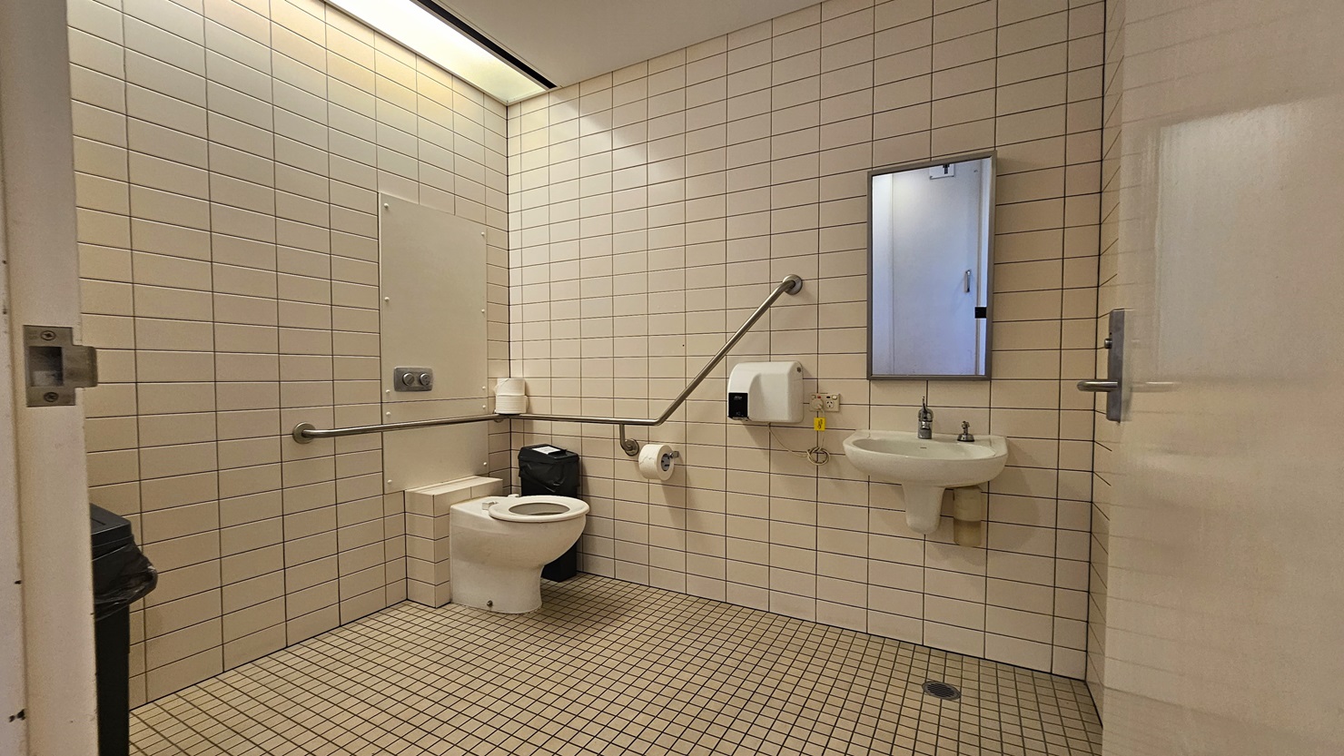 Within the accessible bathroom, a toilet bowl with handrails is situated in the corner. Beside it, a tissue dispenser and a rubbish bin are placed. Adjacent to the toilet, there is a wash basin and a wall-mounted liquid soap dispenser. Another rubbish bin is positioned on the left, near the wall. The walls and floor are adorned with white tiles, while the ceiling also maintains a white colour scheme.
