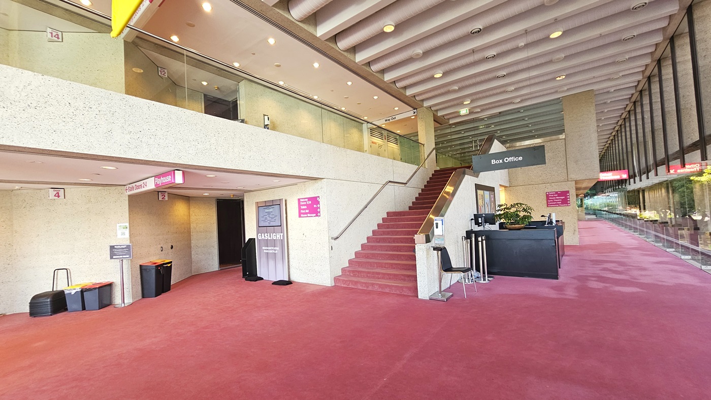In the middle stands a reception desk with a black facade. Beside it are a chair, queue bollard, and sanitising machine. To the left, a staircase with handrails. Wall-mounted signages are visible, along with rubbish bins against the wall. Pink carpet covers the floor, while white walls and ceilings feature large aircon vents and light fixtures. Glass railings adorn the upper floor.