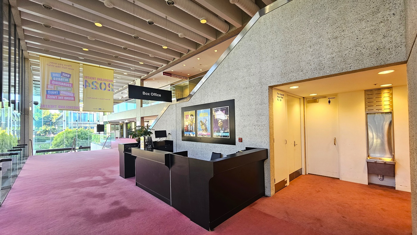 On the left, a black counter showcases colourful theatre posters. Adjacent, a narrow corridor leads to a door with an illuminated 'Box Office' sign above. On the right, floor-to-ceiling grooved concrete walls frame a large doorway covered by a heavy curtain. Throughout, a continuous pink carpet complements austere grey textured walls and exposed ceiling fixtures. Multiple doors on the right lead to the bathroom, with a drinking fountain beside.