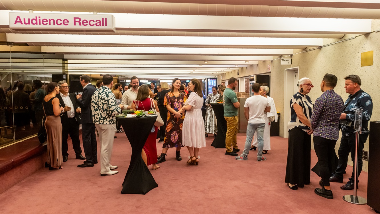 On the left, a group of people converse near a table with refreshments, holding drinks. To the right, more individuals engage in discussion. The background boasts a light pink ceiling with white details and recessed lighting. The floor is carpeted in a darker shade of pink, complementing the venue's colour scheme. Mirrored walls reflect the assembly of attendees. On the right, a section of the bar counter and bathroom doors is visible.