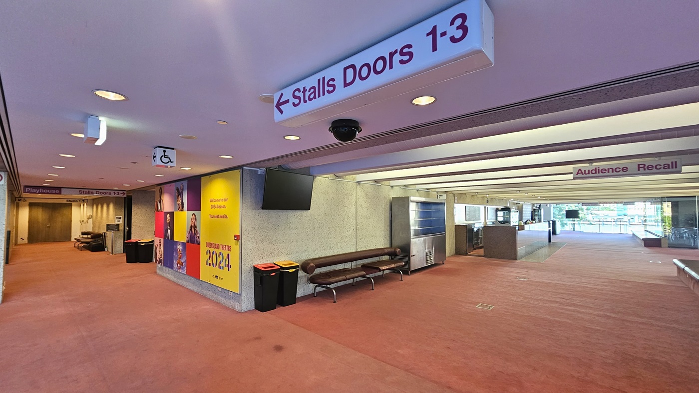 On the left, signage directs to 'Stalls Doors 1-3' above an accessible sign. A yellow banner advertises events. On the right, glass doors lead outside with reflections visible. A counter table and stainless display rack are seen. A seating area with a wooden bench, cushions, and rubbish bins is against the wall. Pink carpet covers the floor, while recessed lighting punctuates the ceiling. An overhead sign reading "Audience Recall" hangs from the ceiling.