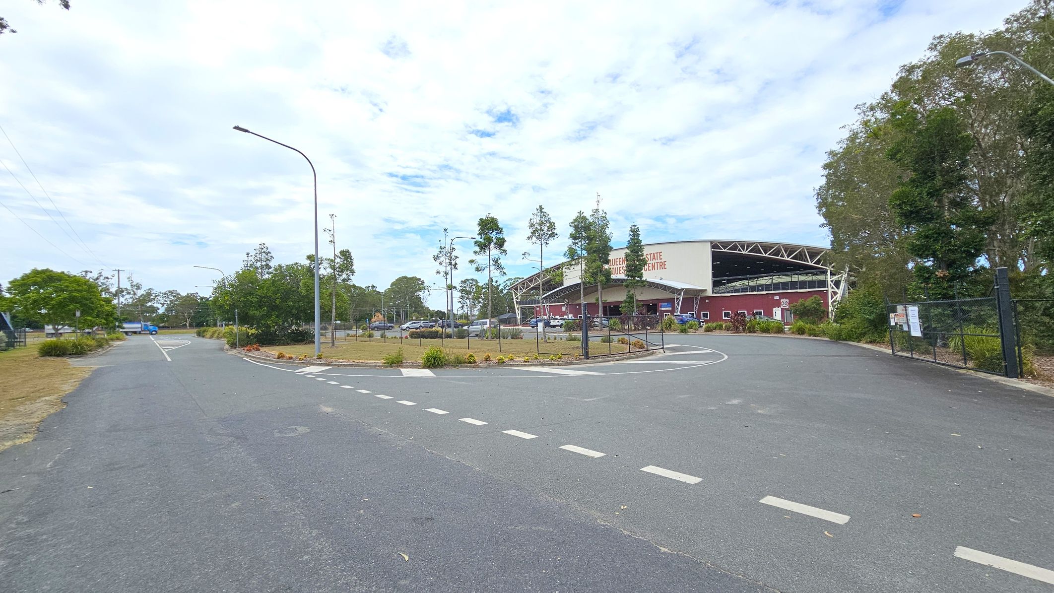 A paved road curves to the right, leading towards a large building with a sign that reads "Queensland State Equestrian Centre." The building has a maroon exterior with a white roof and open sides. On the left, there are trees and a grassy area with a blue vehicle in the distance. The right side features a fence and more trees. Street lamps line the road, and the sky is partly cloudy.