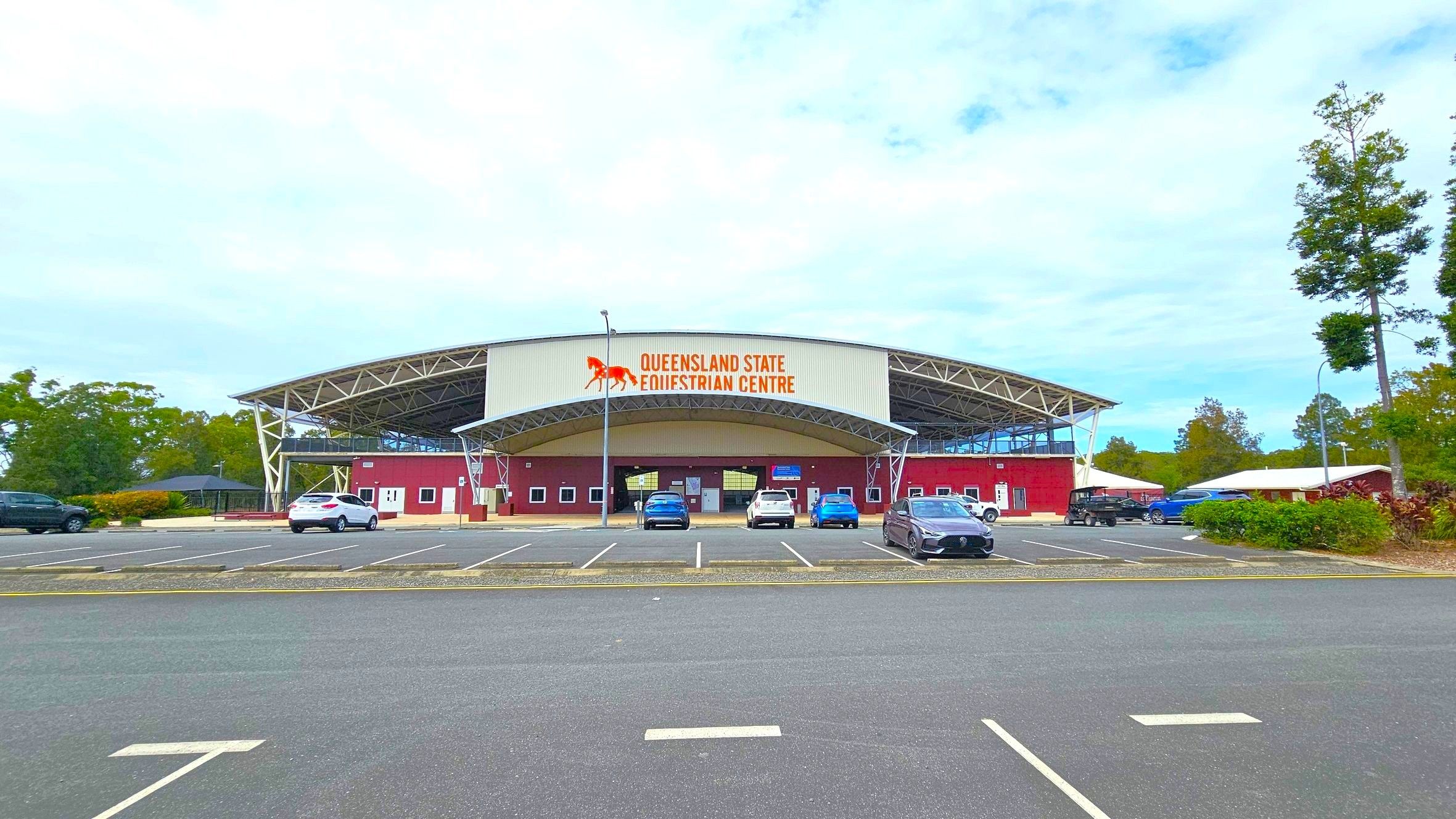 A large equestrian centre with a curved roof and a sign reading "Queensland State Equestrian Centre" in the middle. The building is red with white accents and has multiple small windows. Several cars are parked in front of the building in a spacious car park. Trees and greenery are visible on both sides, with a few tall trees on the right. The foreground shows an asphalt surface with marked parking spaces.
