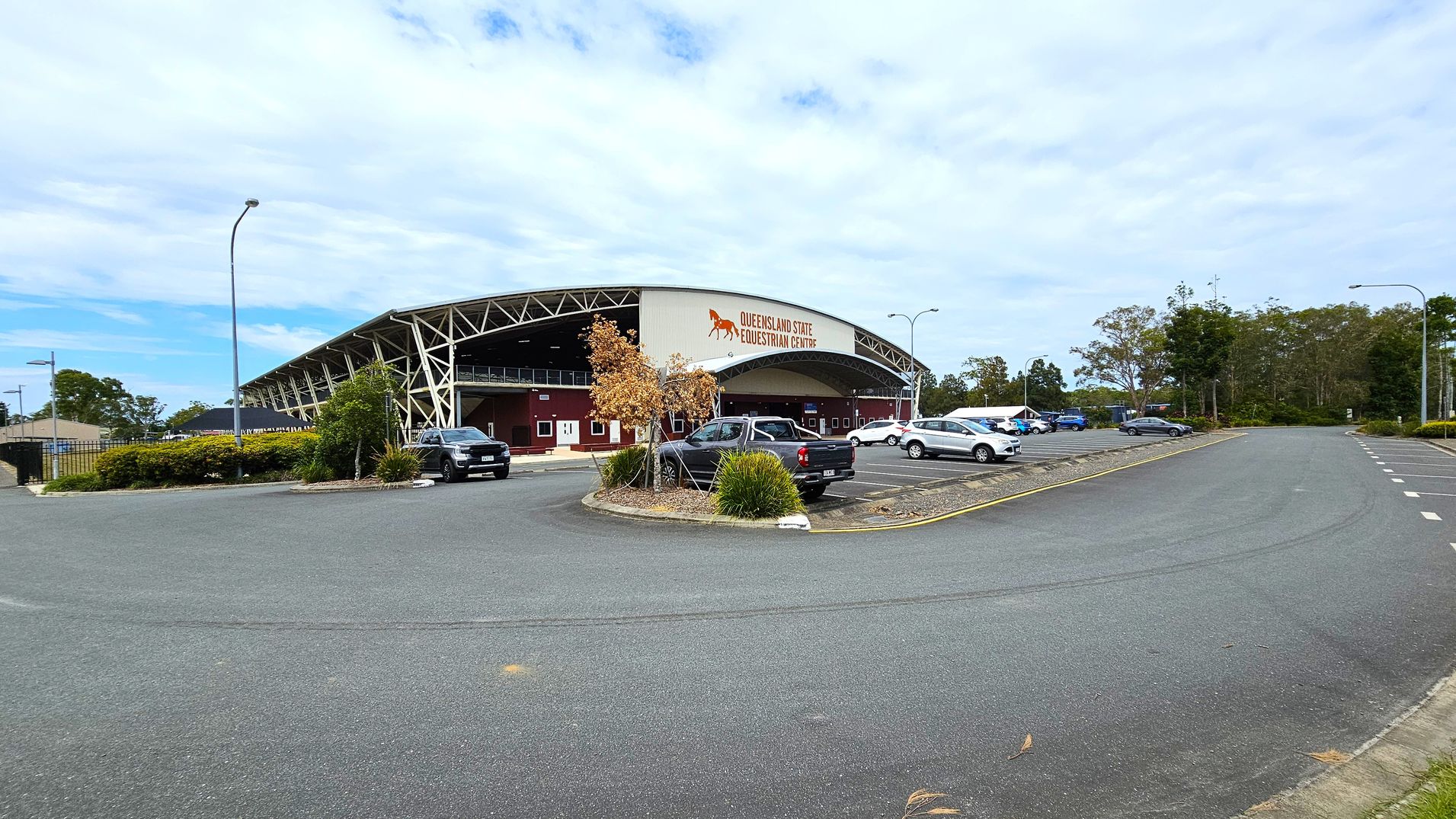 A large, open parking area with several cars is in front of a building marked "Queensland State Equestrian Centre." The building has a curved roof with a metal framework and maroon walls. A few small trees and shrubs are planted around the parking area. The road curves around the parking lot, and there are streetlights along the edges. In the background, there are more trees and greenery.