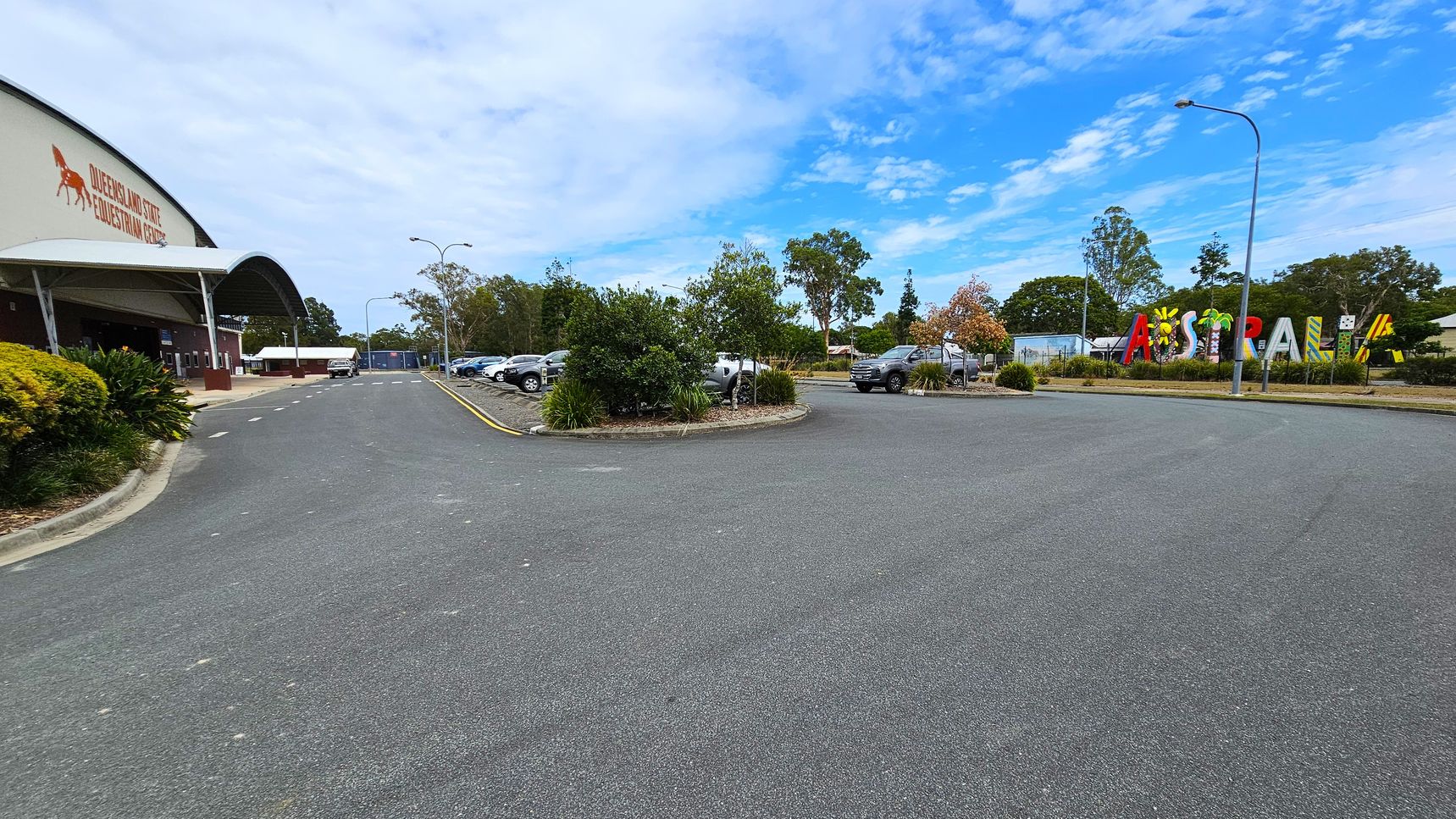 On the left, a large building with a curved roof and signage for the Queensland State Equestrian Centre. The foreground features a wide asphalt road with a few parked cars. On the right, colourful letters spelling "Australia" are visible, surrounded by greenery and trees. The background includes more parked cars and tall trees. Streetlights are positioned along the road.