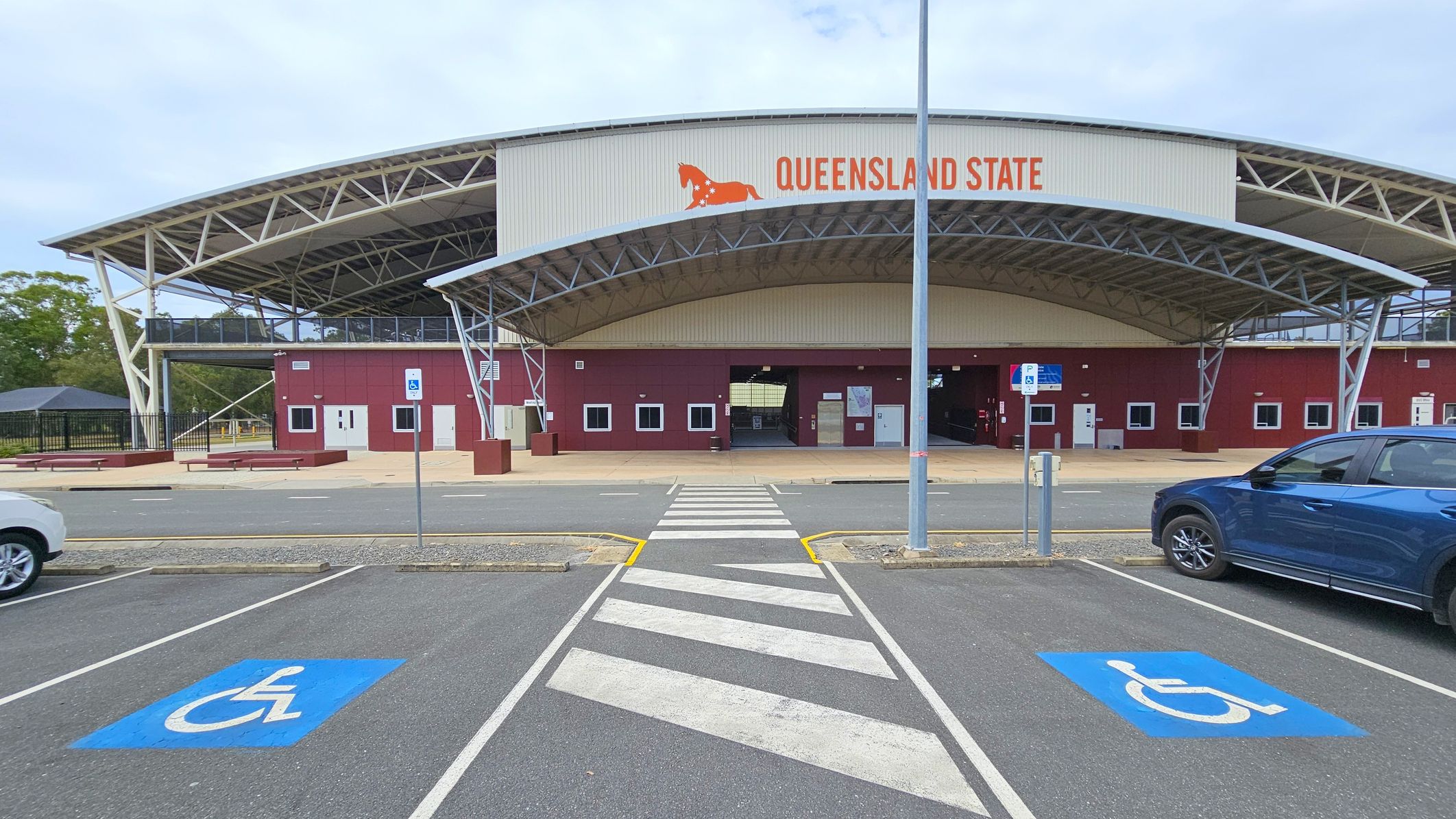 A large building with a curved metal roof and a sign reading "Queensland State" in orange letters. The structure has a maroon facade with several doors and windows. In the foreground, there is a zebra crossing leading to the entrance, flanked by two blue disabled parking spaces marked on the asphalt. A blue car is parked on the right side, and a white car is partially visible on the left. A metal pole stands near the crossing.