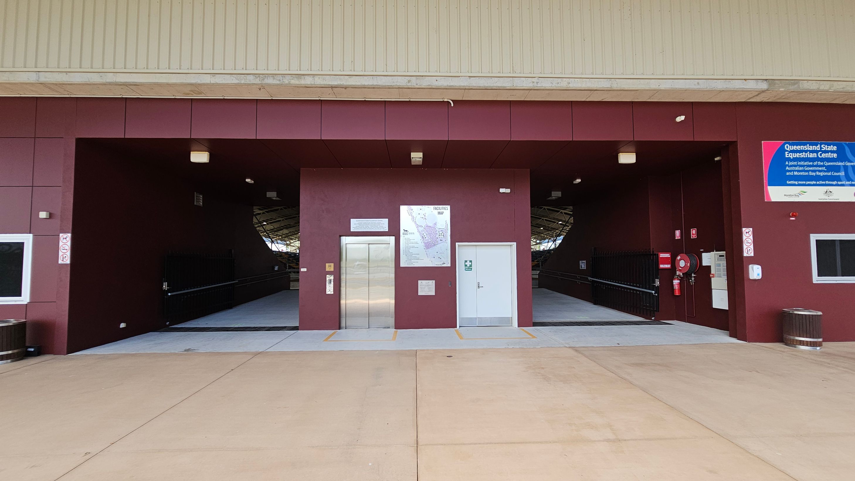 A building entrance with two large open doorways on either side and a central section featuring a lift and a door. The walls are maroon, and there are signs, including a facilities map and a blue sign for the Queensland State Equestrian Centre. The floor is concrete, and there are lights above the entrances. A fire extinguisher and hose reel are mounted on the right wall. The ceiling is light-coloured with visible beams.