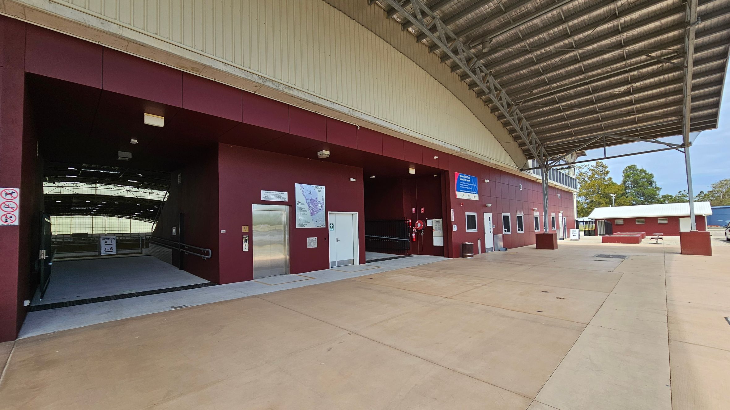 A covered area with a large metal roof and a maroon-coloured building on the left. The building has an entrance with a ramp, an elevator, and several signs, including a map. On the right, there is an open space with a small white-roofed structure and a bench. The ground is paved with light-coloured concrete. Trees are visible in the background.
