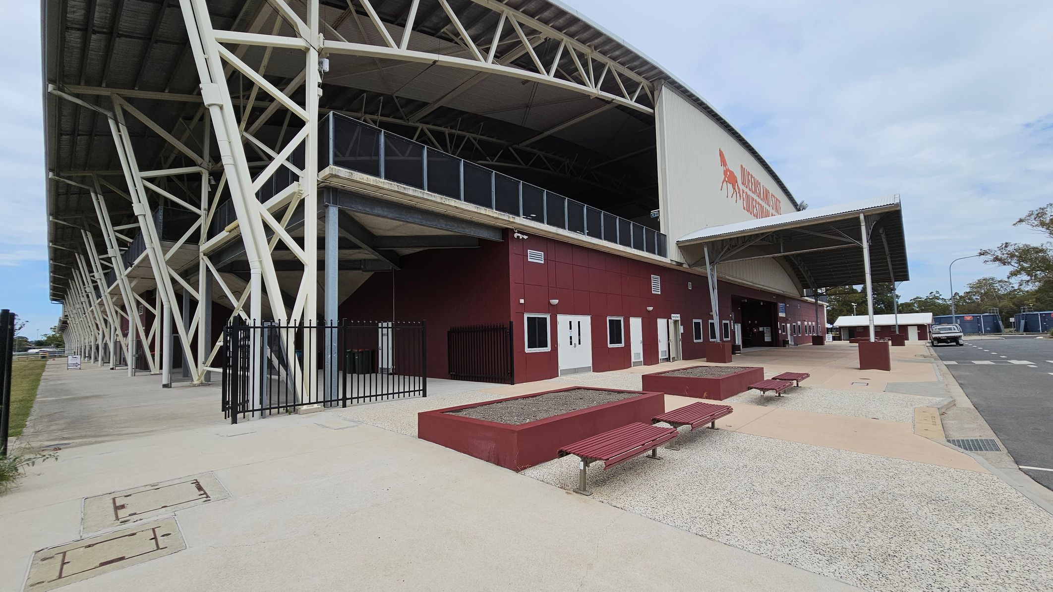 A large stadium structure with a metal roof and white support beams on the left. The building has a maroon facade with several white doors and windows. In the foreground, there are red benches and rectangular planters on a paved area. A black metal fence is visible near the entrance. On the right, a road with a parked vehicle and portable buildings can be seen. Trees are in the background.