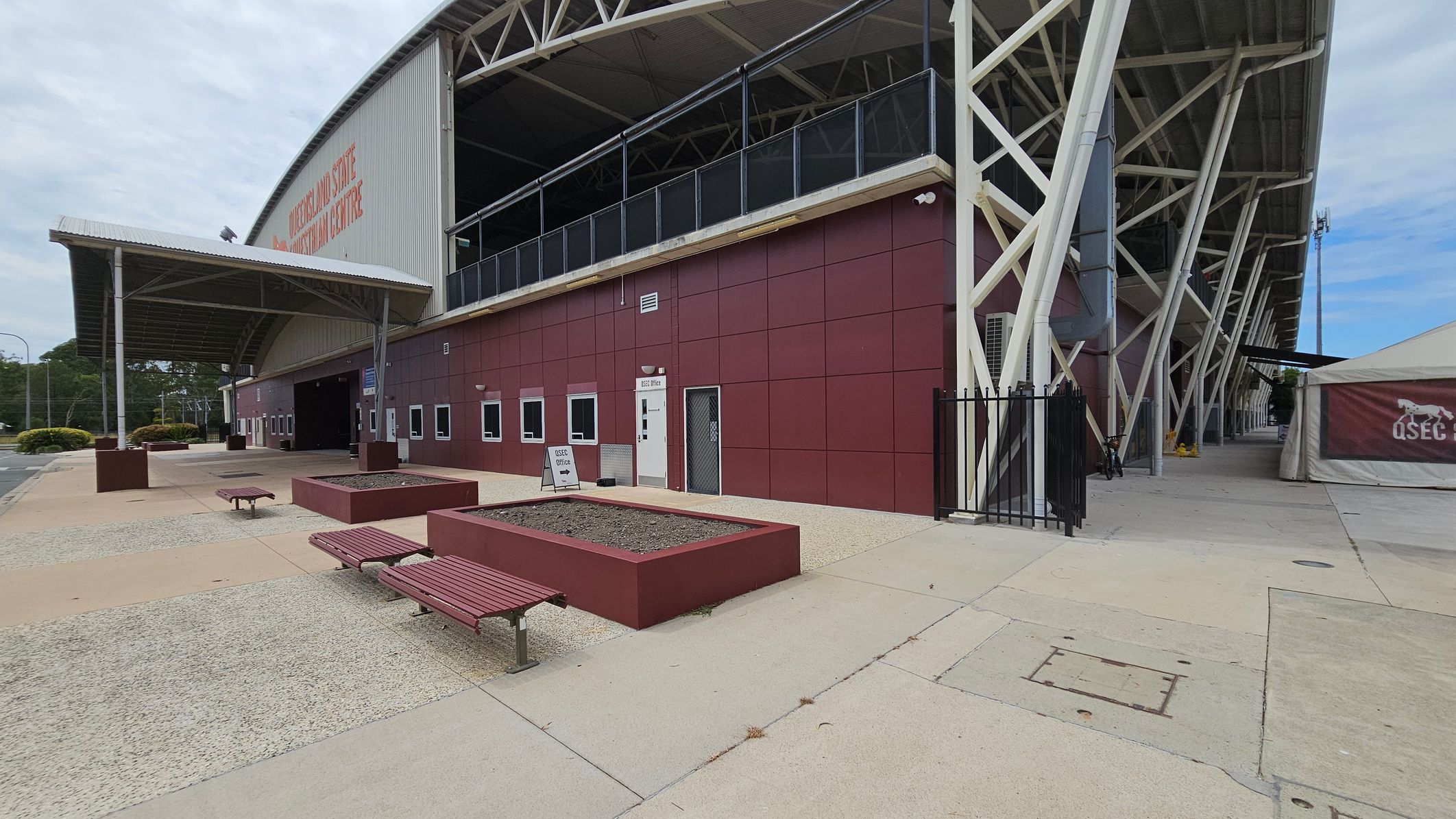 A large building with a curved roof and a sign reading "Queensland State Equestrian Centre" on the side. The structure has a maroon exterior with small windows and a door marked "QSEC Office". In front, there are rectangular planters and benches on a paved area. To the right, a white tent with "QSEC" branding is visible. The building is supported by white structural beams, and the ground is a mix of concrete and textured surfaces.