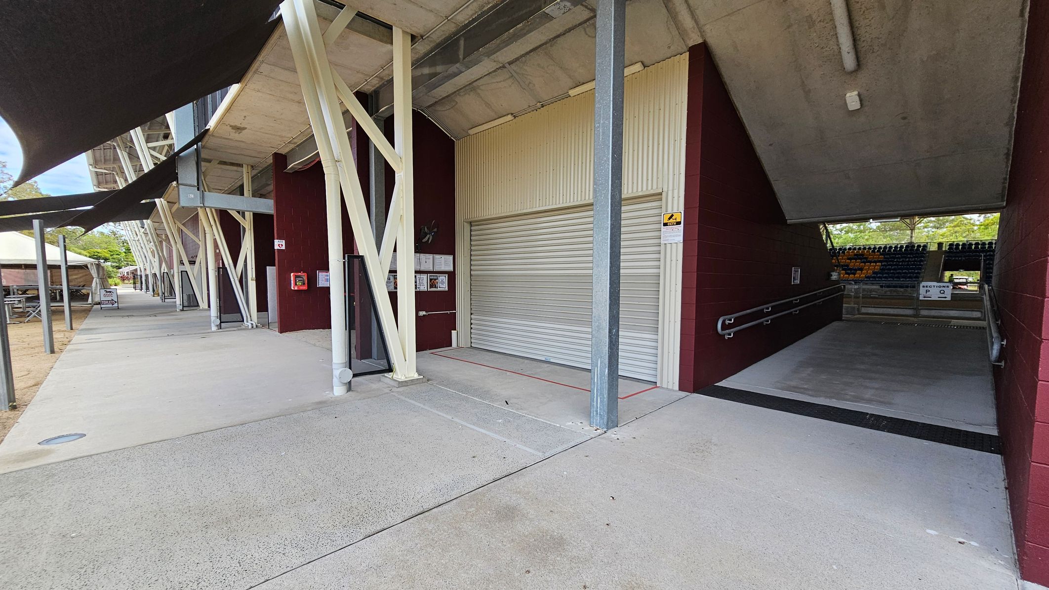 A concrete walkway runs alongside a building with maroon walls and white structural supports. On the right, a closed roller door is set into the wall, next to an entrance ramp leading to a seating area with rows of yellow and black seats. Overhead, a concrete ceiling with a light fixture is visible. To the left, there are outdoor tables under a canopy, and a sign indicating a café. The ground is paved, and the area is shaded by black awnings.