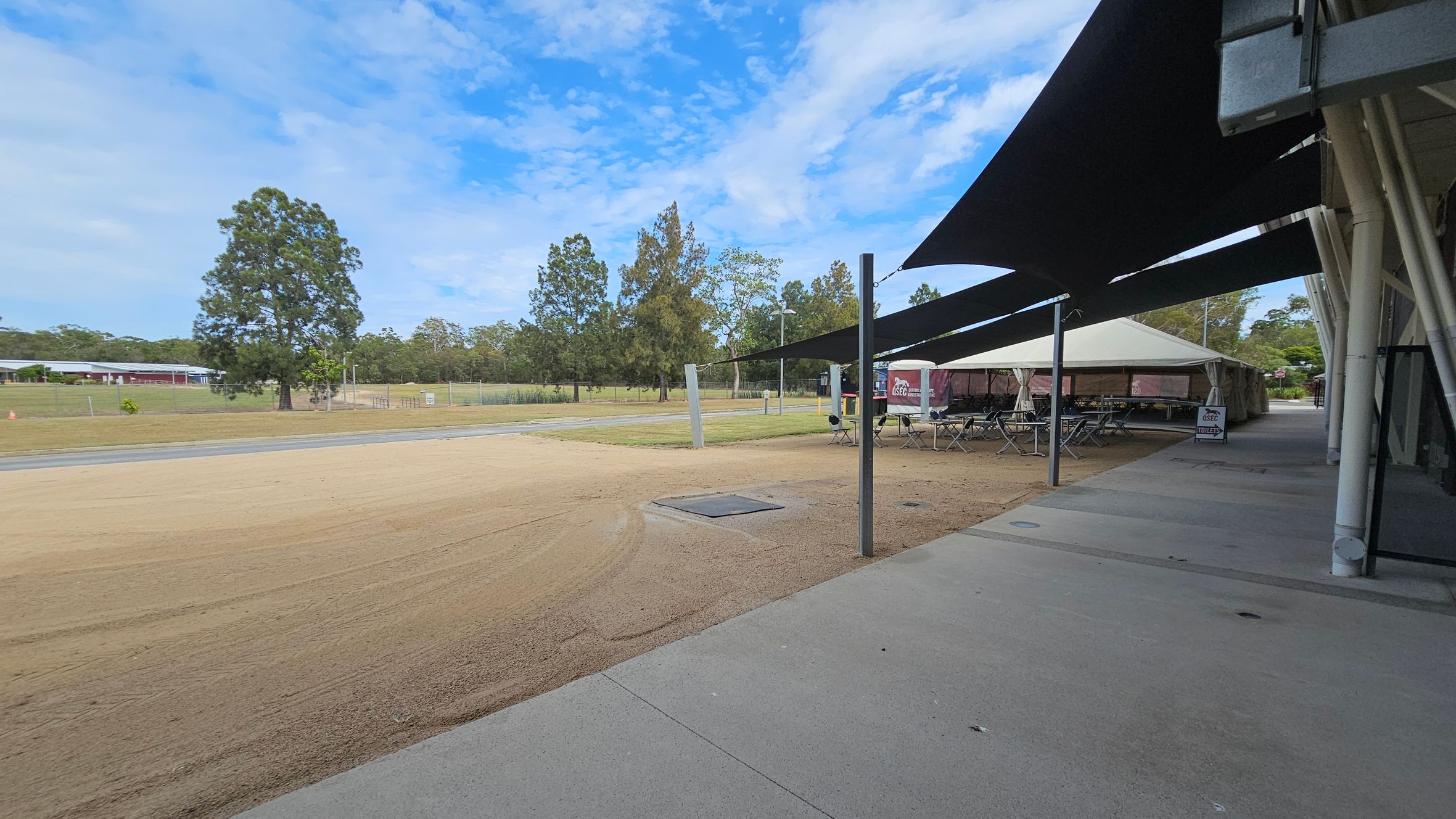 On the left, there is a grassy area with a large tree and a building in the distance. The middle ground features a sandy path and a small paved road. On the right, there is a row of outdoor seating under a large tent with black shade sails extending from a building. The foreground has a concrete walkway, and the background is filled with more trees and greenery.