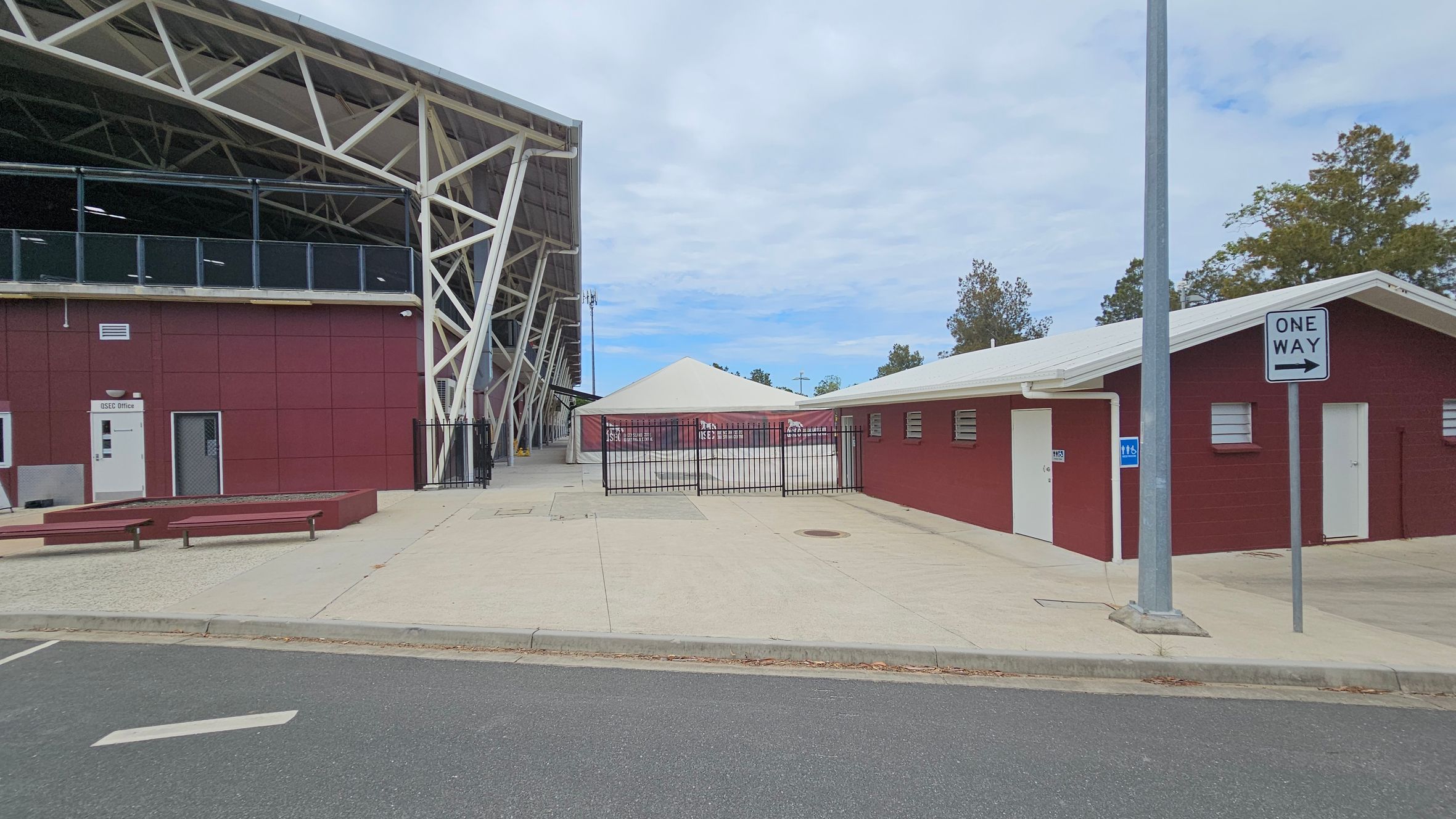 On the left, a large building with a maroon facade and white structural supports. A sign reads "OSHC Office" near a door. In the centre, a fenced area with a white tent-like structure. On the right, a smaller maroon building with a white roof and doors, next to a "One Way" sign on a pole. The ground is paved with a mix of concrete and asphalt. Trees are visible in the background.
