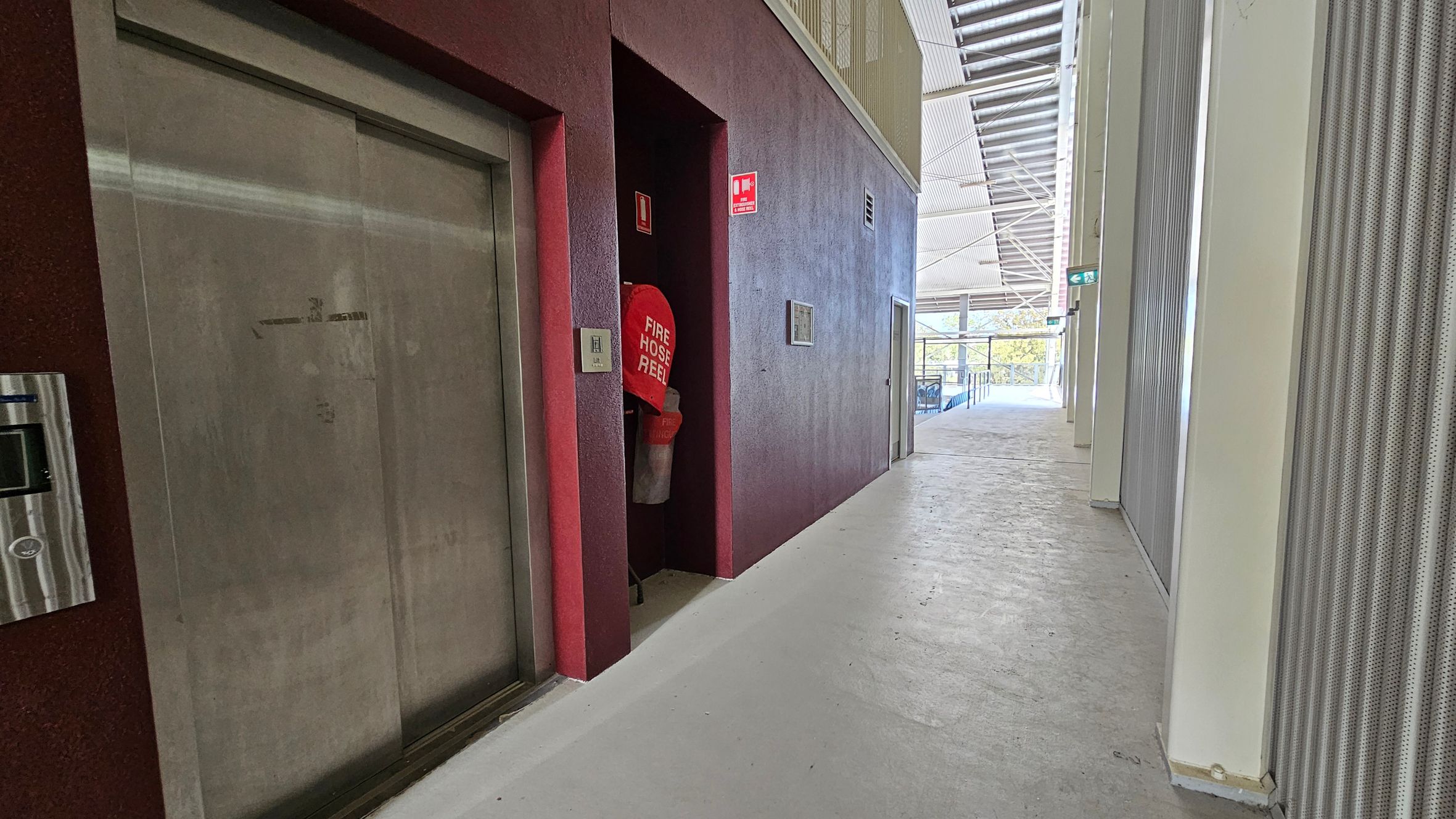 A corridor with a concrete floor and a metal ceiling. On the left, there's a closed lift with a metallic door and a fire hose reel in a recessed area. A red fire safety sign is visible above. The right side has a series of vertical, perforated panels. In the background, large windows allow natural light in, and there's a view of outdoor greenery. An exit sign is visible above a door at the end of the corridor.