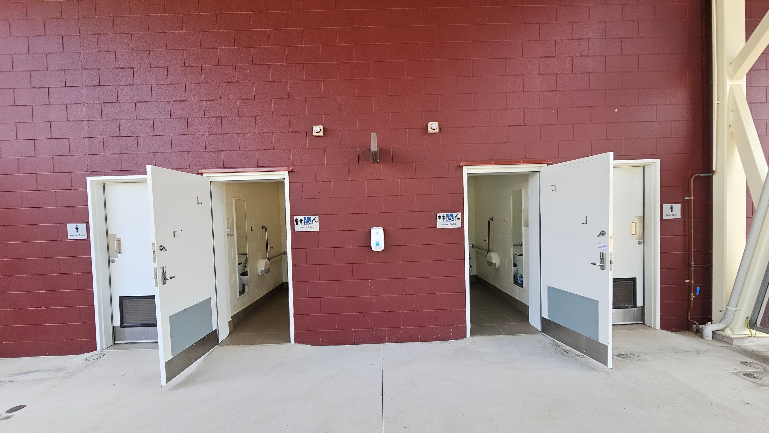 A red brick wall with two open doors leading to accessible toilets. Each door has a sign indicating unisex and accessible facilities. The floor is concrete, and there is a hand sanitiser dispenser mounted on the wall between the doors. Inside, the toilets have white walls and are equipped with grab bars. Pipes and structural beams are visible on the right side of the image.