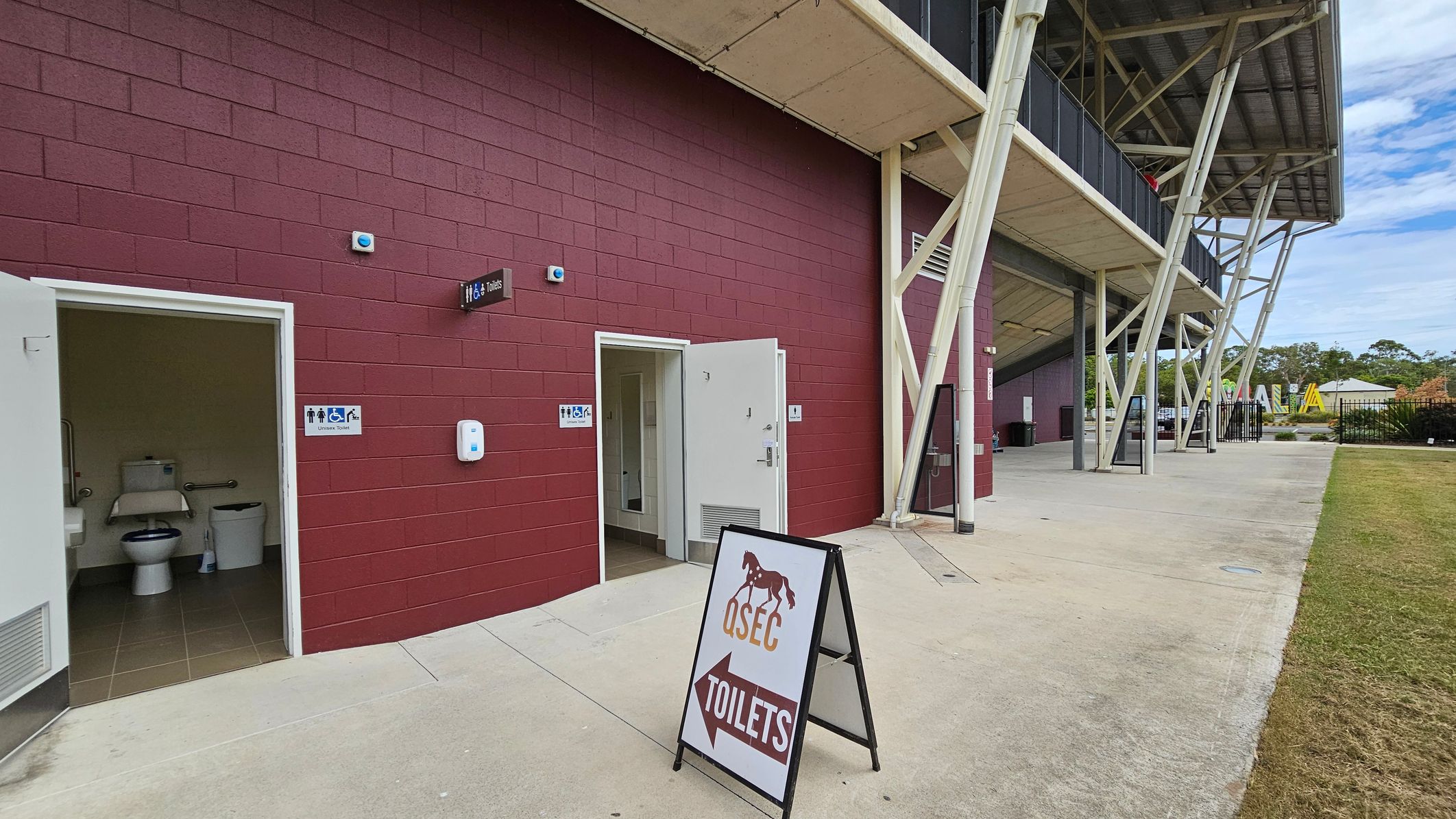 A red brick building with two open doors leading to toilets. Signs indicate male, female, and accessible facilities. A freestanding sign with a horse graphic reads "QSEC Toilets" with an arrow. The building has a metal roof supported by angled beams. The ground is concrete, and there's a grassy area to the right. In the background, there are trees and a structure with a roof.