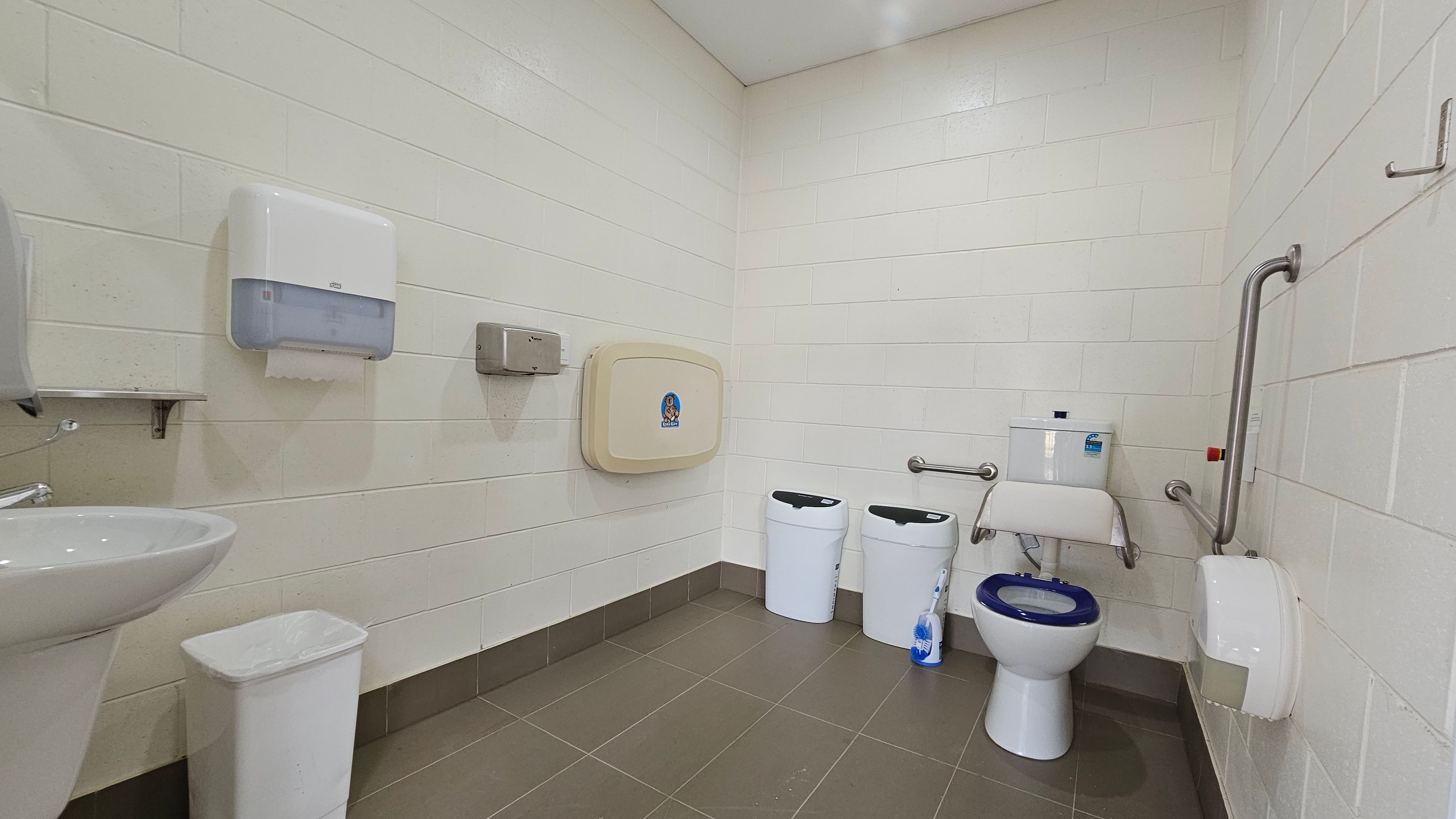 A public restroom with white tiled walls and a brown-tiled floor. On the left, there is a sink with a white bin beneath it, and a paper towel dispenser above. In the centre, a baby changing station is mounted on the wall. To the right, there are two white bins, a toilet with a blue seat, and a toilet brush. Stainless steel grab bars are installed around the toilet, and a toilet paper dispenser is mounted on the wall. The ceiling is plain and light-coloured.