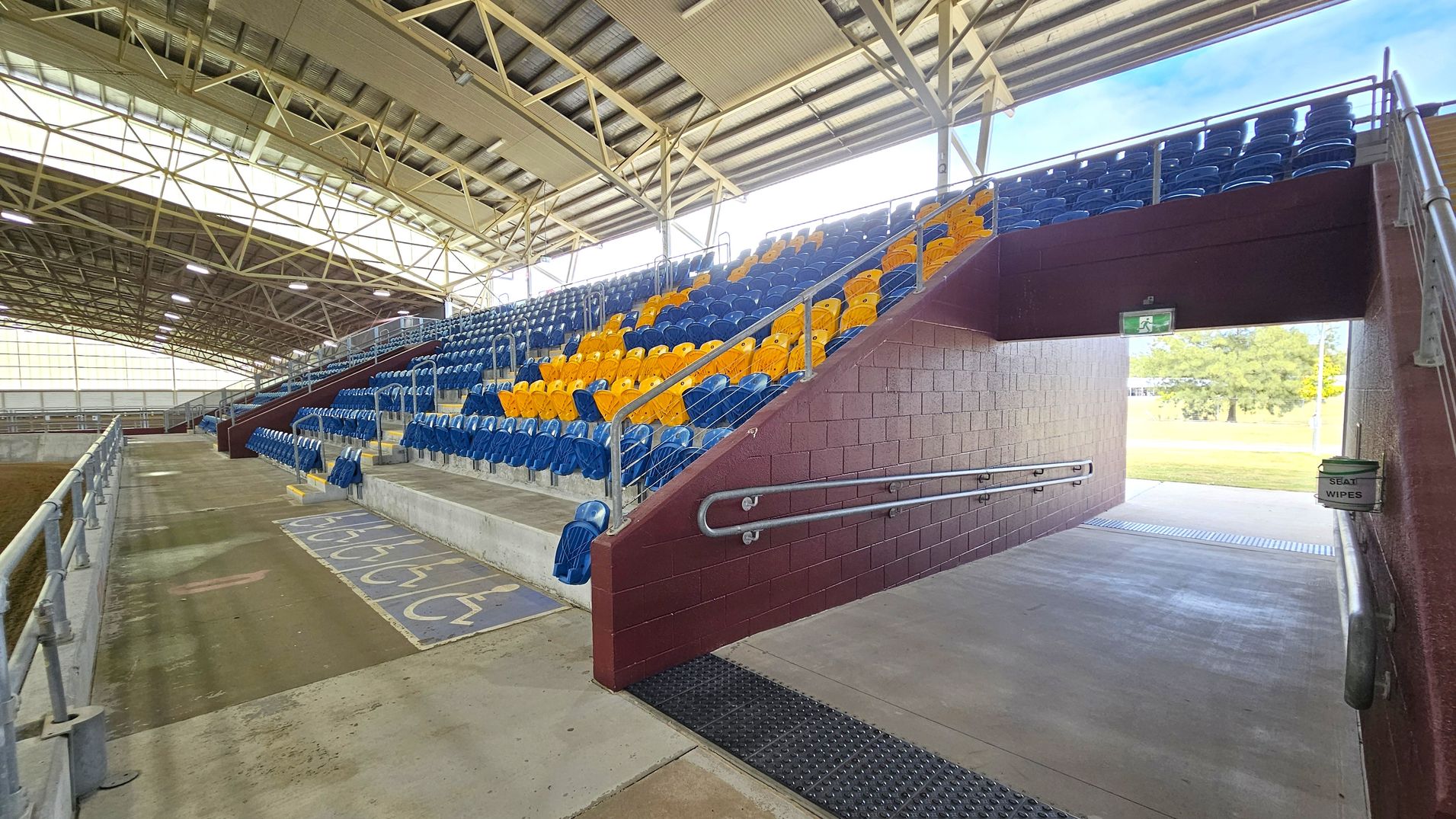 Indoor sports arena with tiered seating on the right, featuring blue and yellow plastic seats. A maroon brick wall with a passageway leads outside, where a grassy area is visible. A sign for seat wipes is attached to the wall. The left side has a concrete floor with wheelchair markings and a metal railing. The ceiling is supported by a metal framework with large windows allowing natural light.