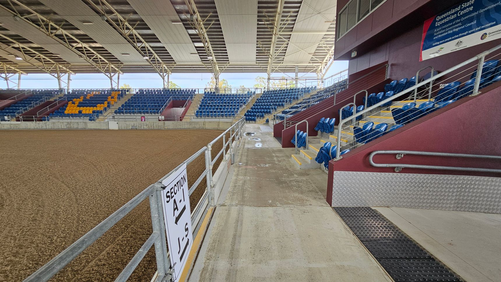 An indoor equestrian arena with a dirt floor on the left. On the right, there are rows of blue seats with some yellow seats forming a pattern. A sign indicates "Section A-J." The seating area is elevated with metal railings and steps. The ceiling is supported by a metal framework with large panels. A large sign is visible on the upper right wall. The arena is well-lit with natural light coming through the open sides.