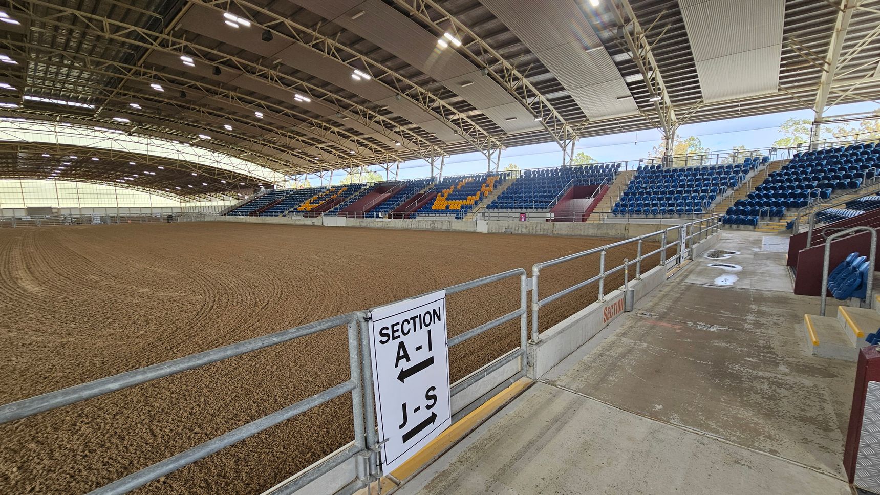 An indoor arena with a large dirt floor, surrounded by tiered seating. On the right, blue and yellow seats are arranged in sections, with a concrete walkway leading up to them. A sign on a metal railing indicates seating sections A-I and J-S. The ceiling is high with exposed beams and lighting fixtures. The left side shows more seating and the expansive dirt area. The arena is well-lit with natural light coming through translucent panels.