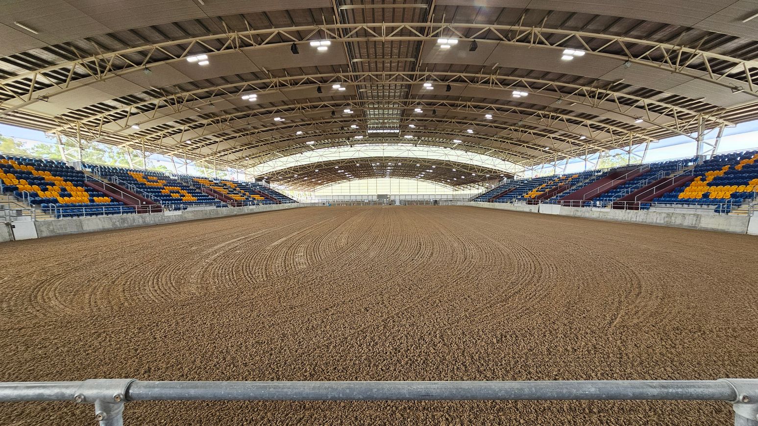 An indoor arena with a dirt floor featuring circular patterns. On both sides, there are tiered seating areas with blue and yellow seats. The ceiling is supported by a metal framework with multiple lights. The background shows a large, translucent wall allowing natural light to enter. A metal railing is visible in the foreground.