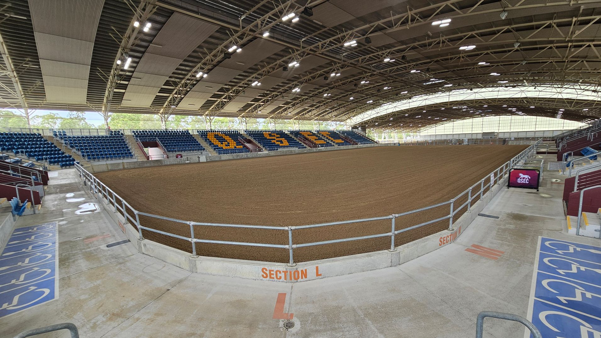 An indoor arena with a large dirt floor in the centre, surrounded by tiered seating. The seats on the left and right are blue, with some sections displaying yellow letters. The ceiling is arched with exposed beams and lighting fixtures. The floor is concrete with marked sections, including a designated area for accessibility. A railing separates the seating from the arena floor. A digital screen on the right displays a logo. The background shows trees visible through the open sides.