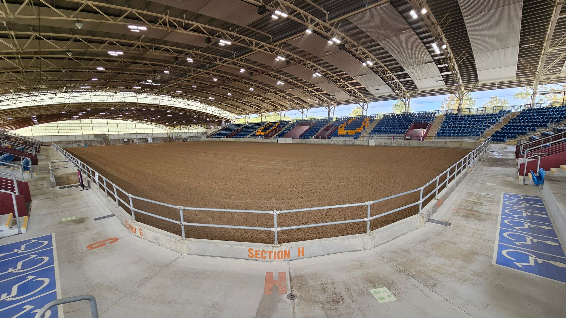 An indoor arena with a large dirt floor in the centre, surrounded by tiered seating. The seating on the right features blue chairs with some yellow ones forming patterns. The ceiling is high with exposed beams and lighting fixtures. The foreground shows a concrete floor with marked sections for accessibility, including wheelchair symbols. A railing separates the seating area from the arena floor, and "Section H" is marked on the barrier. The background includes large windows allowing natural light.