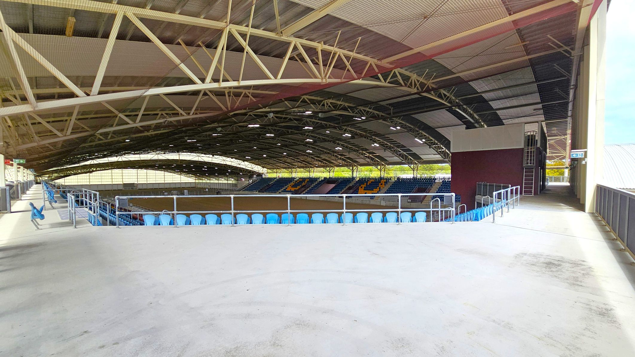 An indoor sports arena with a large, curved roof supported by metal trusses. The seating area features rows of blue chairs, with a section of yellow seats forming a pattern. The floor is concrete, and there are railings along the seating area. On the right, a maroon wall with a ladder leads to an upper level. The ceiling is a mix of metal and translucent panels, allowing natural light to filter through. A walkway runs along the right side, leading to an exit.