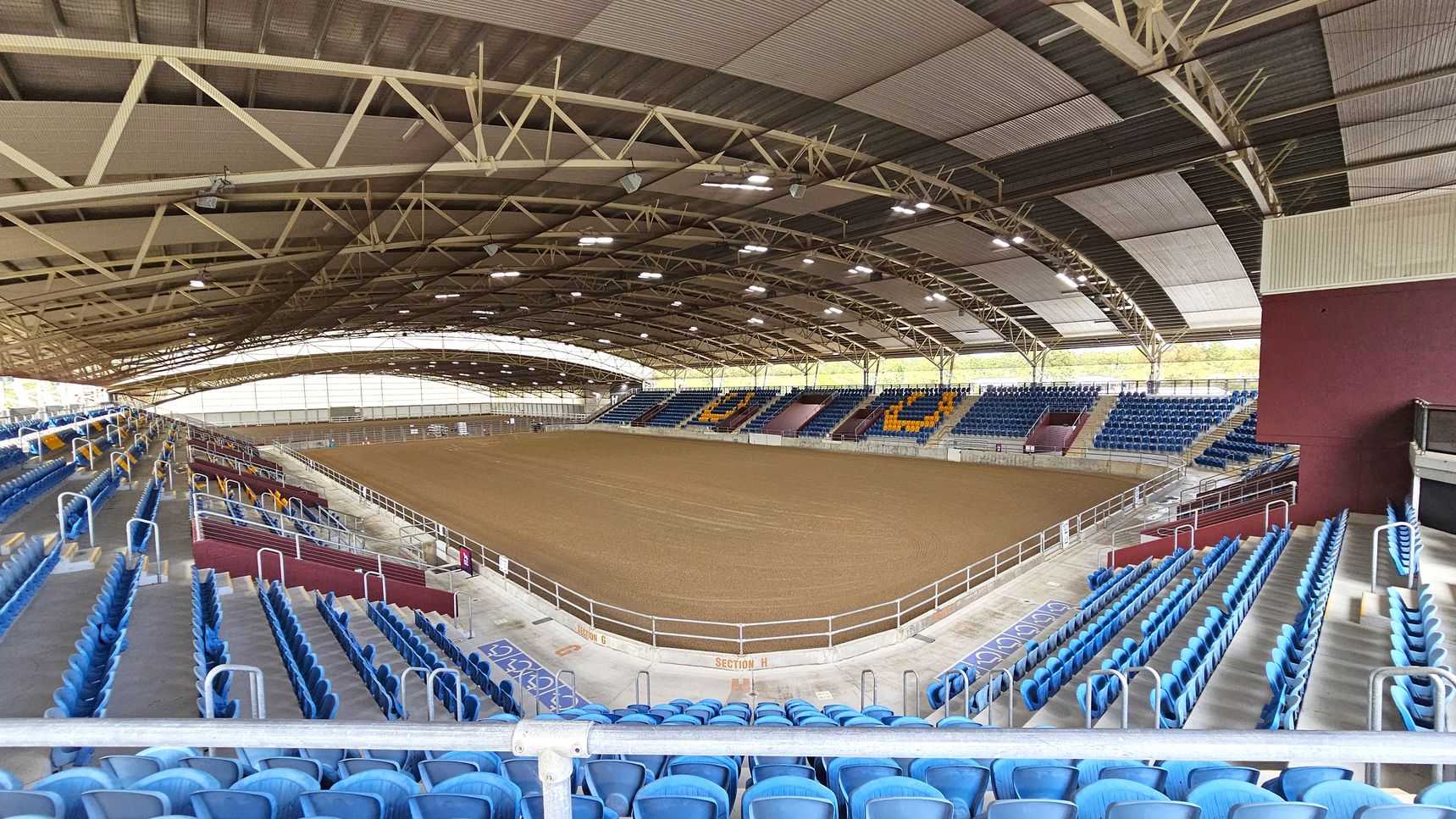 An indoor arena with a large dirt floor in the centre, surrounded by tiered seating. The seats are predominantly blue, with some sections featuring yellow seats forming patterns. The ceiling is high with a metal framework and multiple lights. The left side has more seating with handrails, while the right side shows a maroon wall and additional seating. The arena is spacious and well-lit, with a clear view of the entire area.