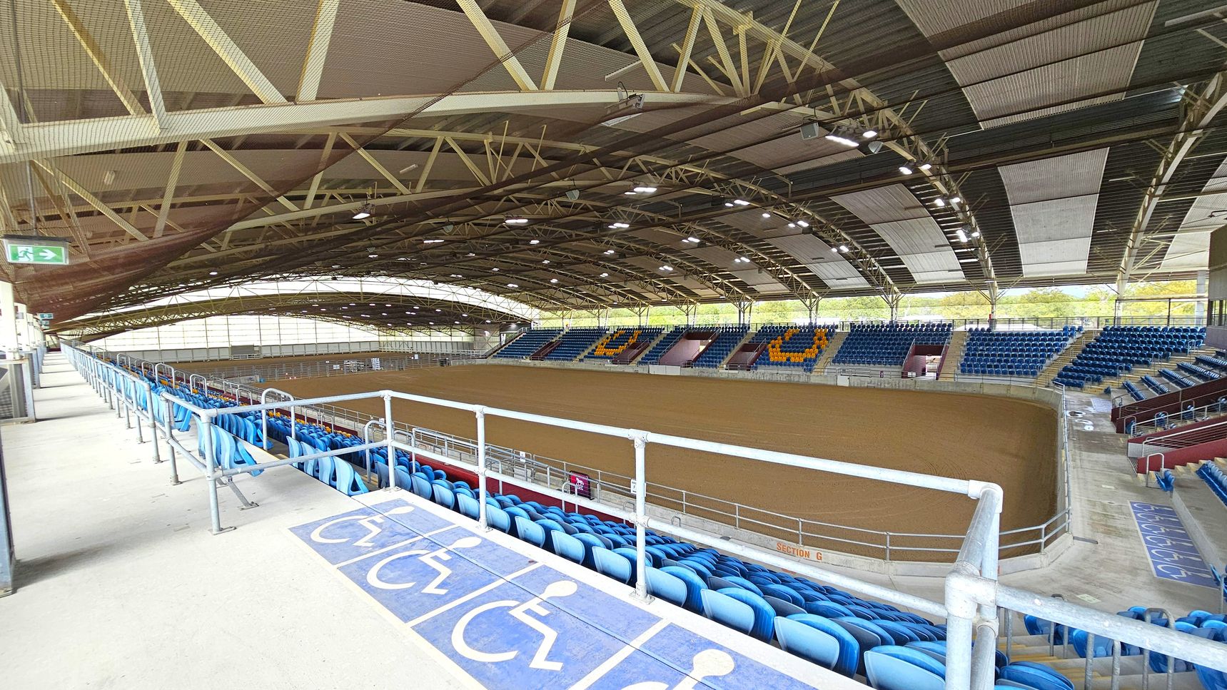 An indoor arena with a large, open dirt floor in the centre. Surrounding the arena are tiered seating areas with blue chairs, some arranged to form patterns. The foreground shows a section with accessible seating marked by wheelchair symbols on the floor. Overhead, a high, arched ceiling with exposed beams and lighting fixtures. An exit sign is visible on the left. The arena is well-lit with natural light coming through large windows on the far side.