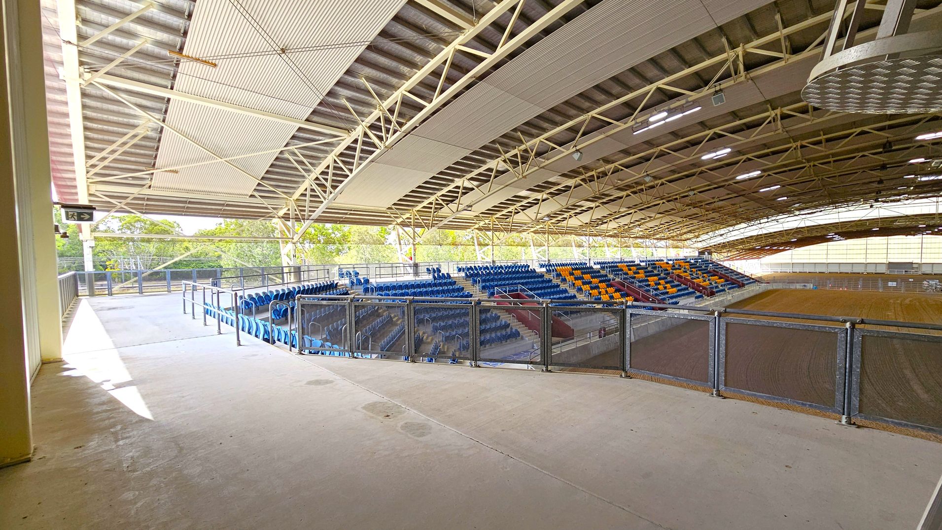 An indoor arena with a large, open space. On the right, there are tiered seating areas with blue and yellow seats. The seating is surrounded by metal railings. The floor is a mix of concrete and dirt, suggesting a multipurpose use. The ceiling is high with exposed beams and metal roofing. On the left, there is a walkway with a railing, leading to an exit sign. The background shows trees visible through the open sides of the structure.