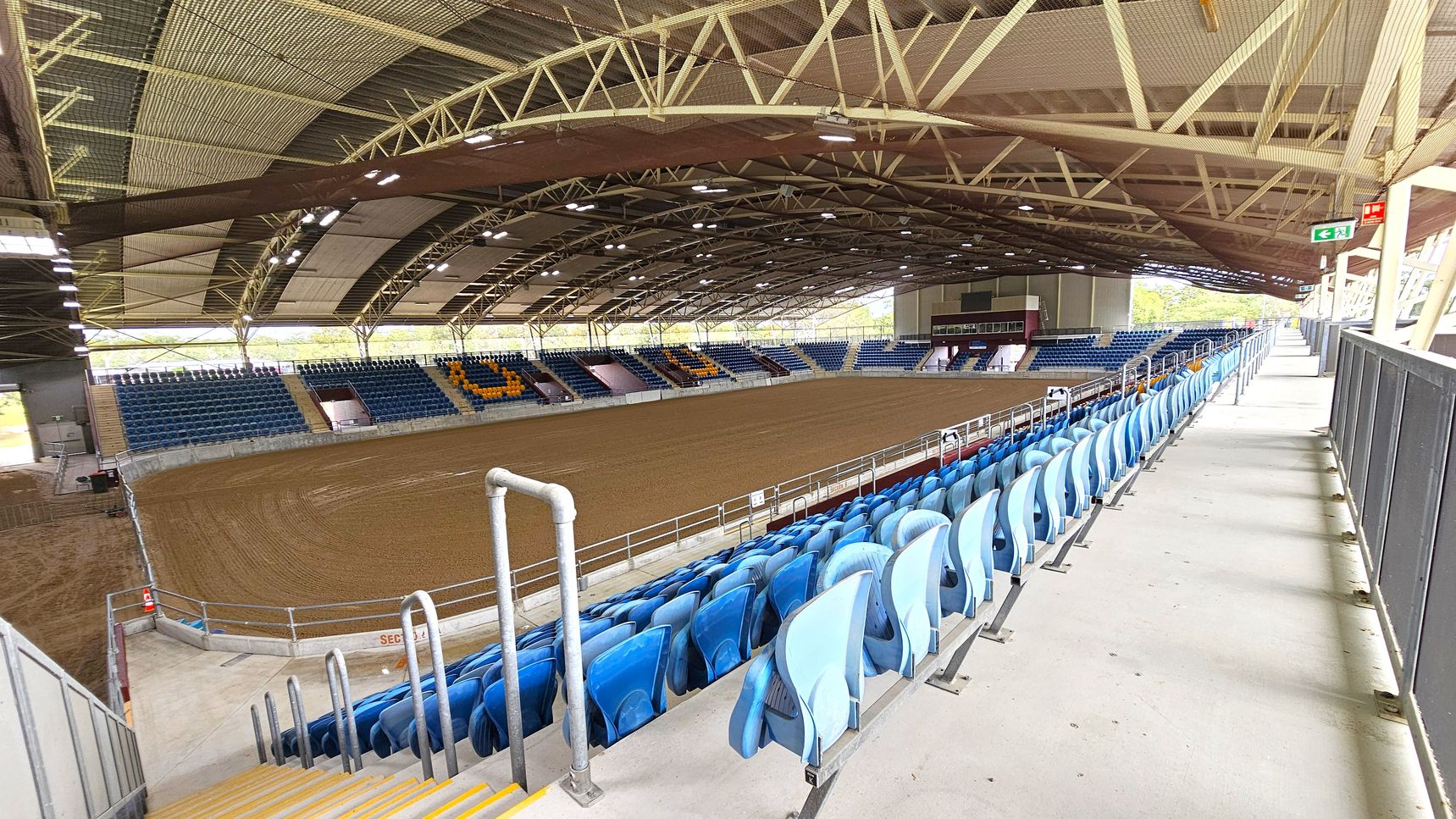An indoor arena with a large dirt floor in the centre, surrounded by tiered seating. On the right, there are rows of blue plastic seats with metal railings and a concrete walkway. The left side features more seating, with some sections in blue and others in yellow. The ceiling is supported by a network of metal beams and is equipped with lighting. An exit sign is visible on the right. The arena is spacious and well-lit, with natural light coming through openings on the sides.