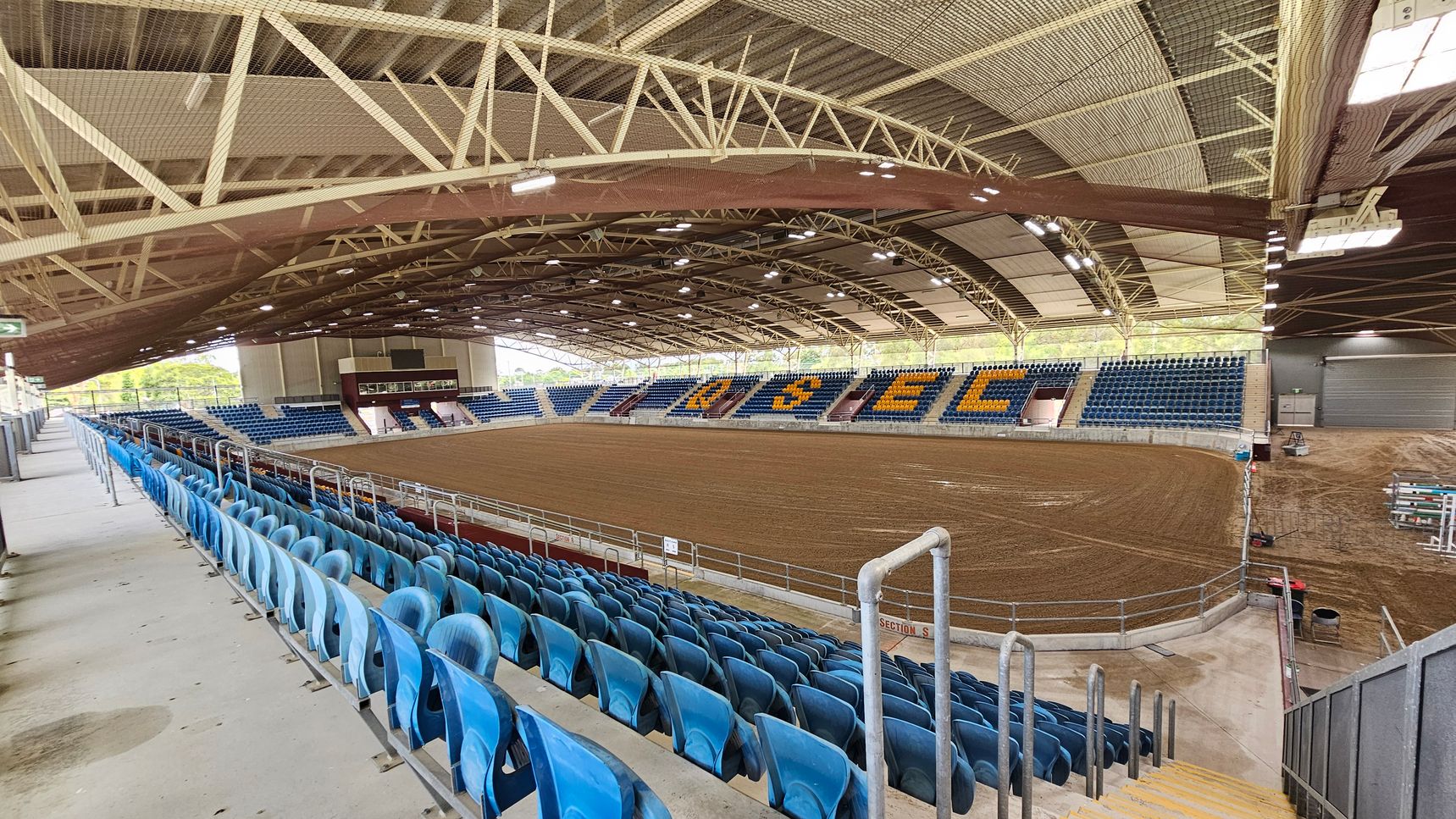 An indoor arena with a large dirt floor, surrounded by tiered seating. The seats are primarily blue, with some sections spelling out letters in yellow. The ceiling is supported by a network of metal beams and features overhead lighting. On the left, there are rows of blue seats leading down to the arena floor. The right side shows more seating and a section with equipment. The background includes a scoreboard and more seating. The floor is concrete around the seating area.