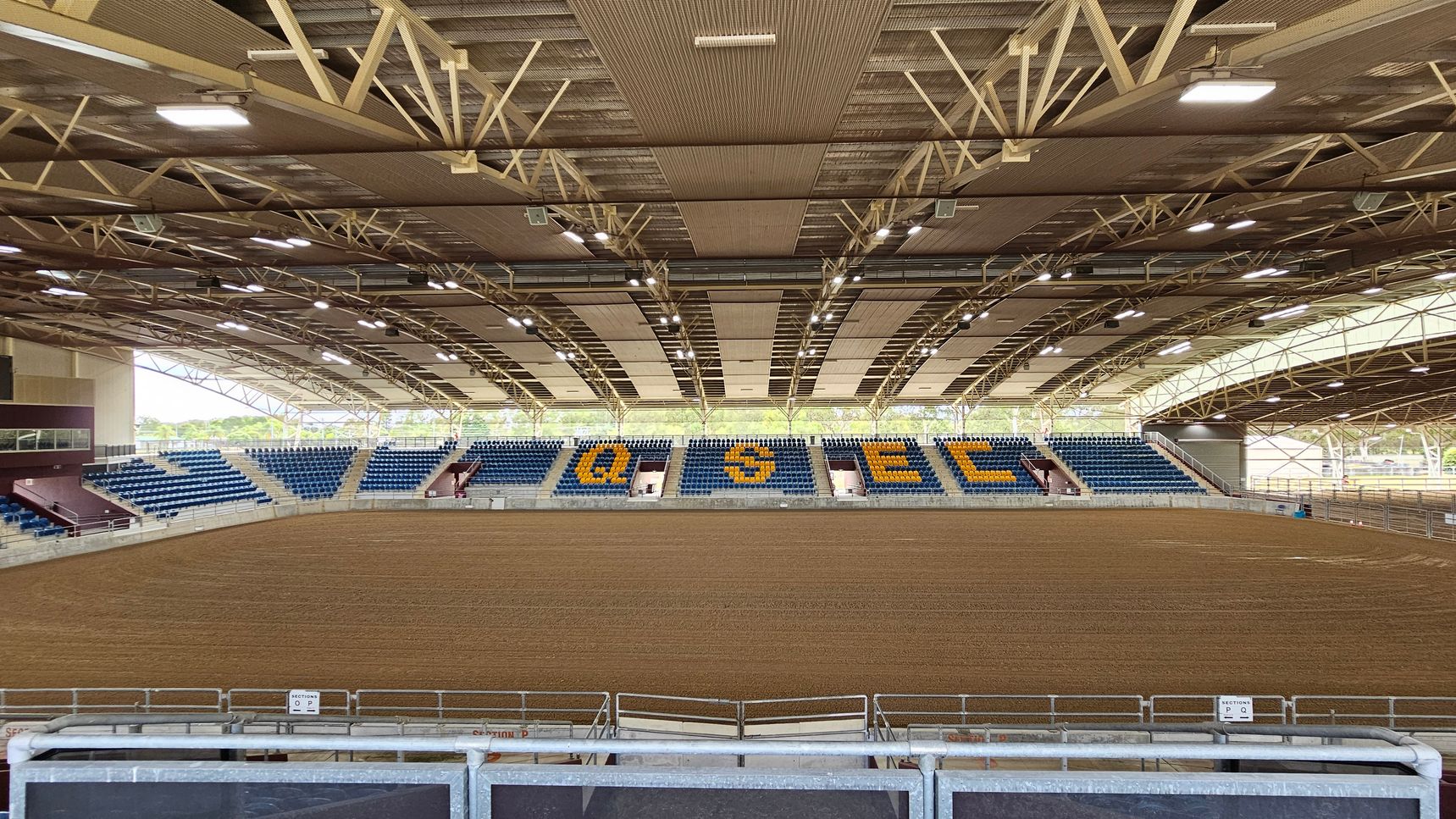 An indoor arena with a large dirt floor in the foreground. The seating area in the background features blue and yellow seats, with letters "QSEC" prominently displayed. The ceiling is supported by a network of metal beams and is equipped with multiple lights. The left and right sides have additional seating and open areas, with some natural light coming through the sides. The arena is enclosed with metal railings.